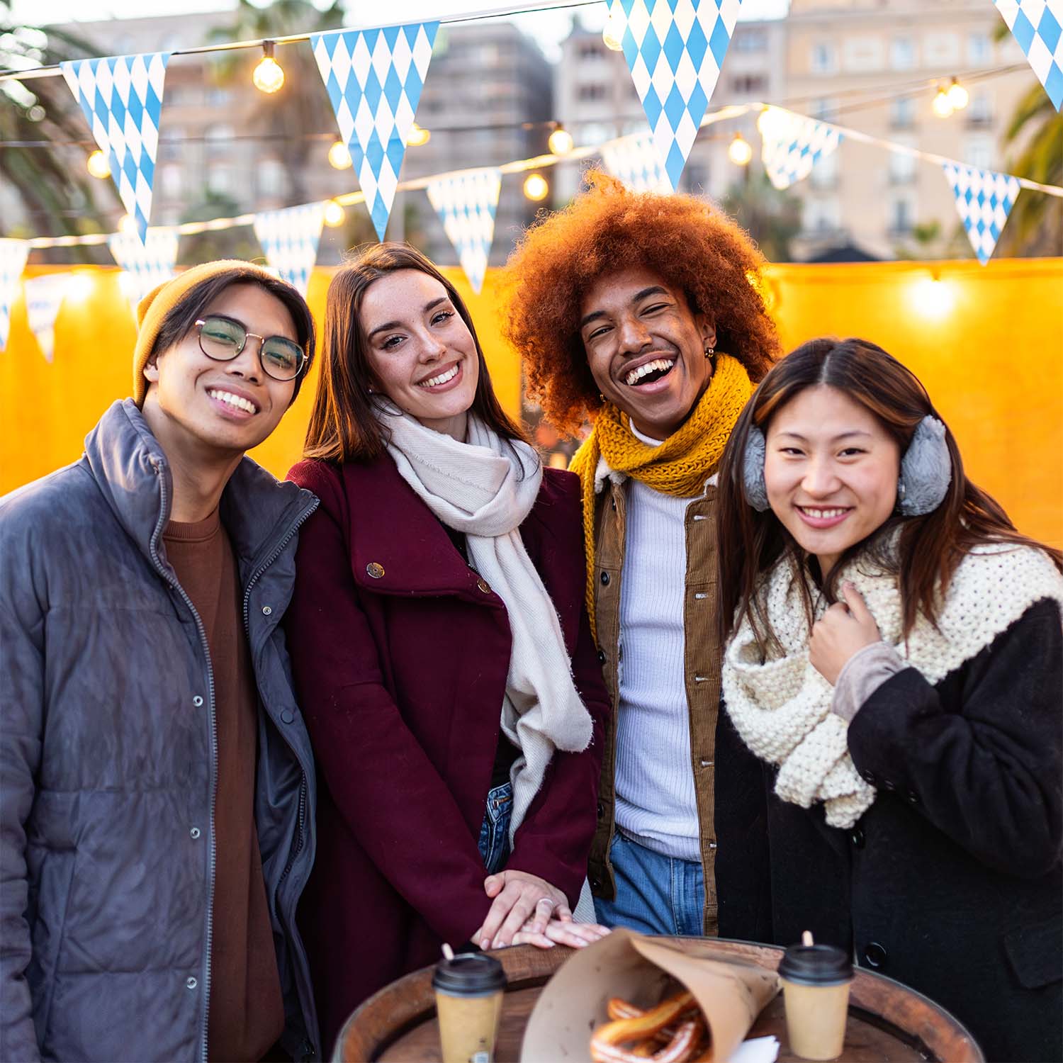Vier jongeren met winterkleding lachen samen bij een tafel met pretzels en koffiebekers buiten, versierd met blauwe en witte vlaggen.