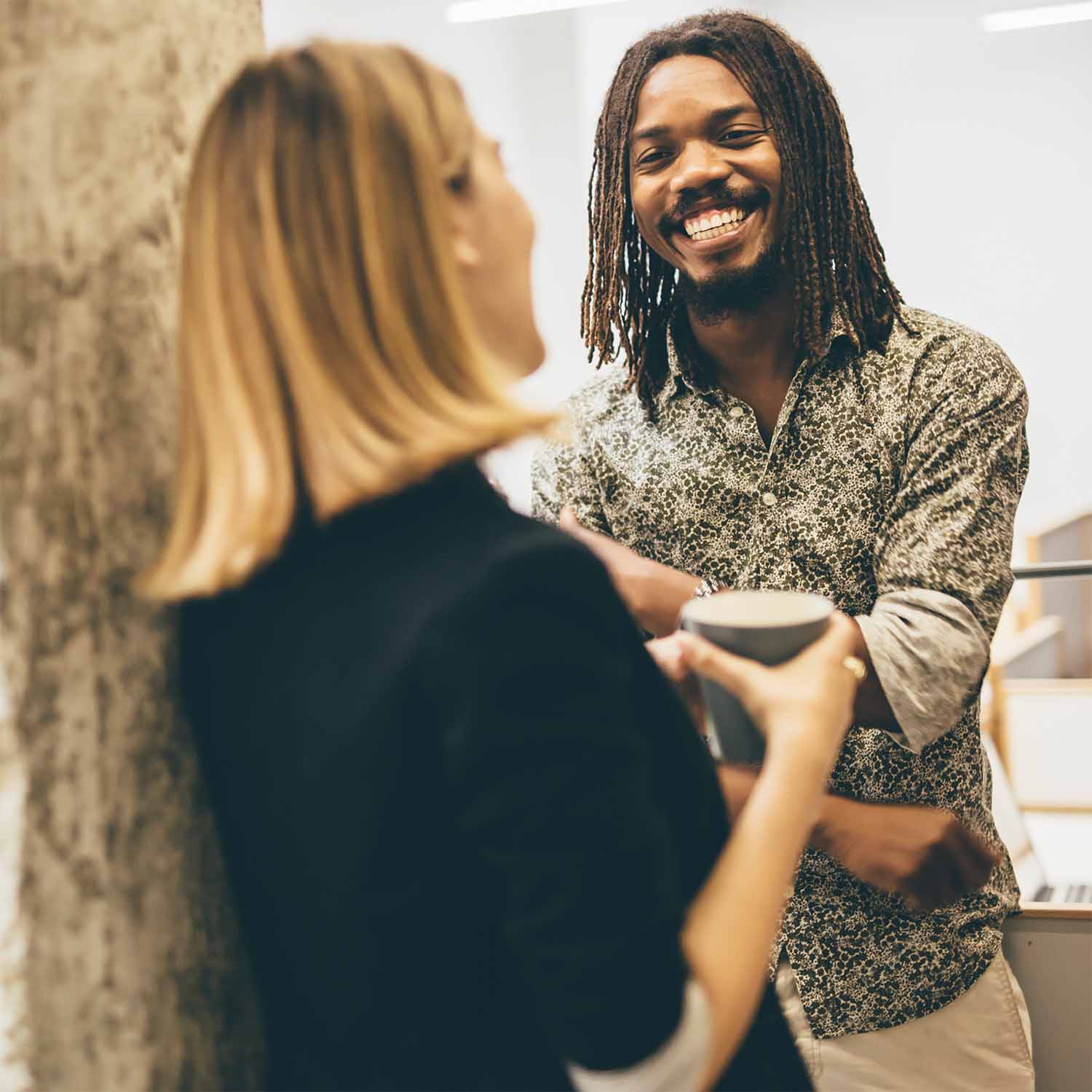 Twee mensen converseren ontspannen, waarbij een glimlachende man met dreadlocks een vrouw met blond haar en een beker aankijkt.