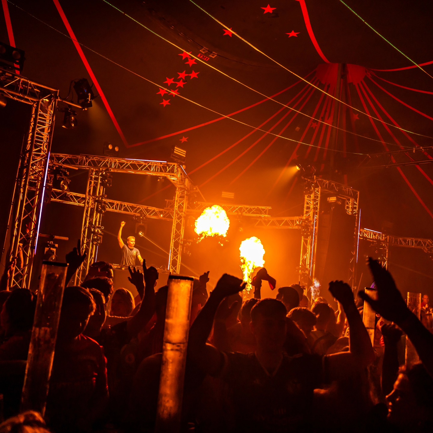 Volle danceclub met publiek dat danst, een DJ die met zijn arm omhoog staat en twee vlammen die uit de stage komen.