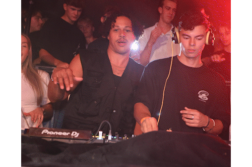 Twee jonge mannen bedienen een DJ-mixer in een club met mensen die op de achtergrond dansen.