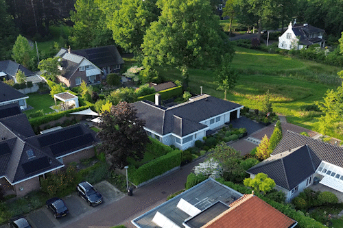 Luchtfoto van een rustige woonwijk met groene tuinen, meerdere huizen en geparkeerde auto's langs de straat.
