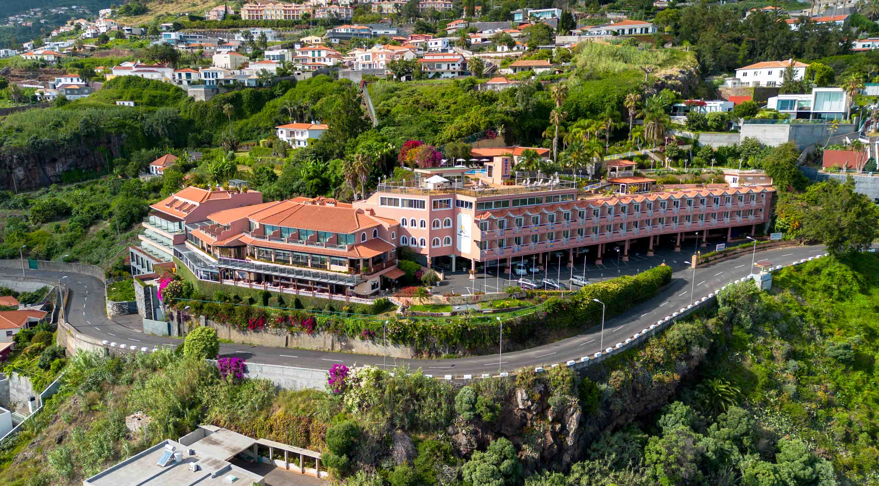 Luchtfoto van een groot resort met terracotta daken, omgeven door groen en heuvelachtig landschap met een kronkelende weg ervoor.