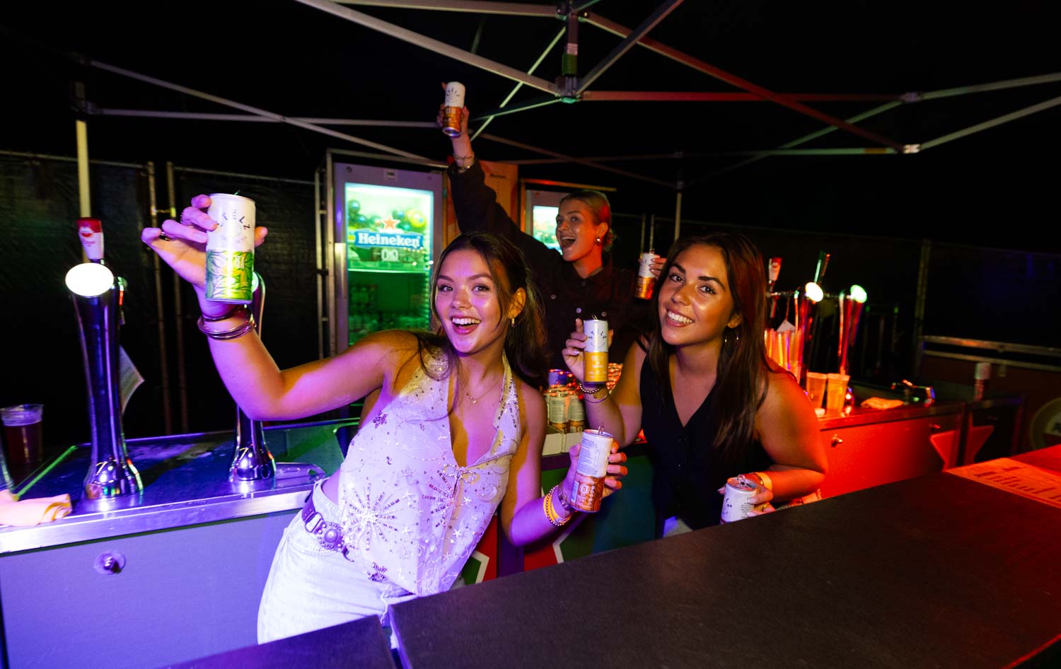 Drie jonge vrouwen vieren feest bij een bar en houden blikken drank omhoog, lachend en genietend van het moment.