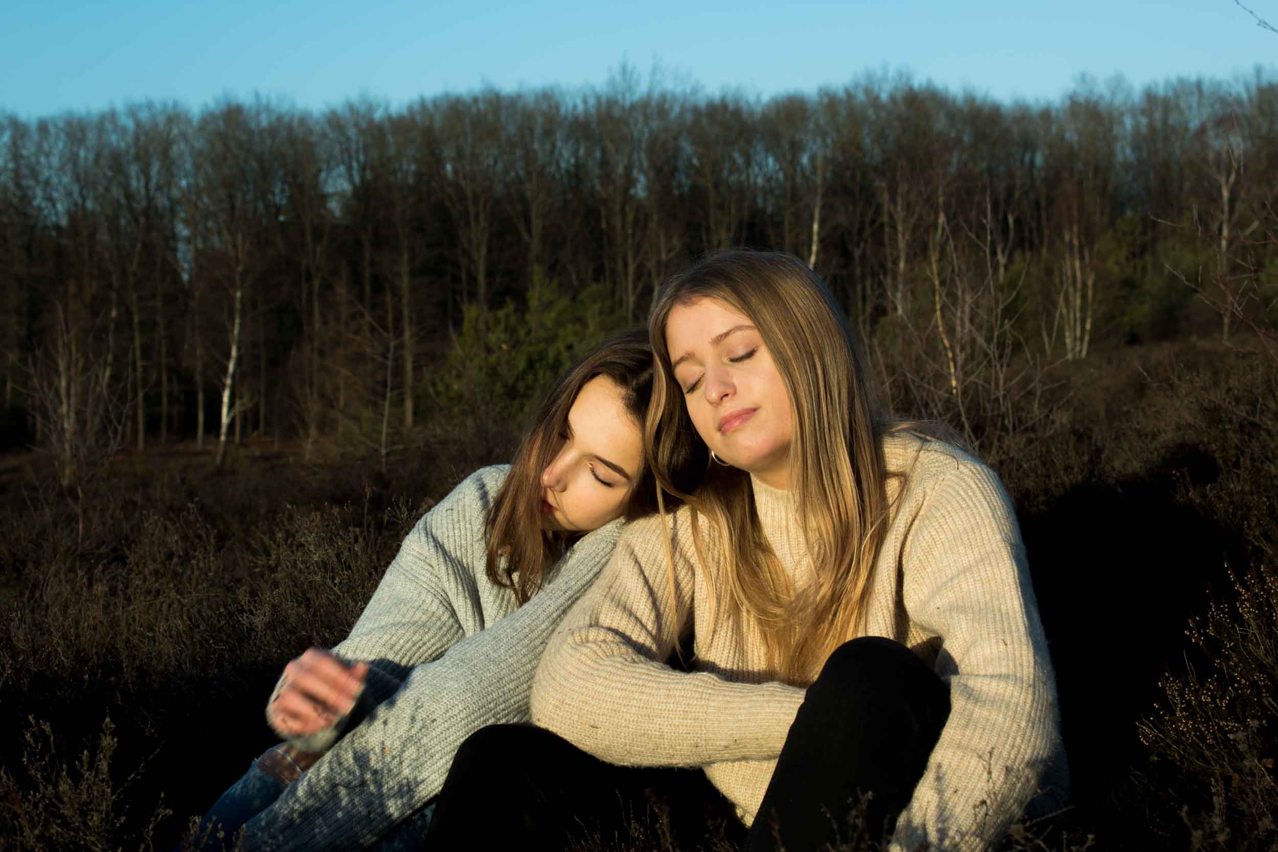 Twee jonge vrouwen met gebreide truien zitten dicht bij elkaar buiten, de ene leunt met gesloten ogen op de schouder van de ander die ook haar ogen gesloten heeft.