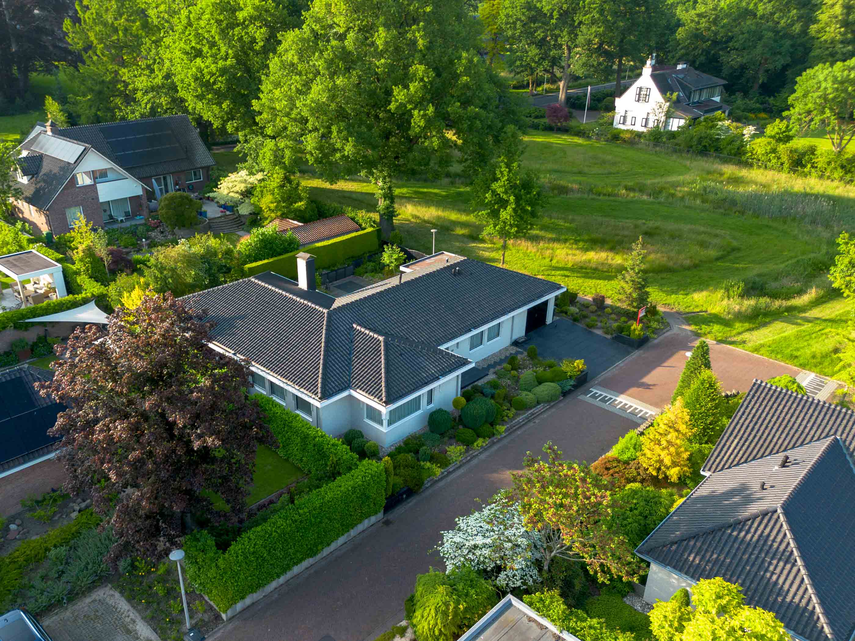 Luchtfoto van een woonwijk met huizen omgeven door groene bomen en tuinen, zonnige dag.