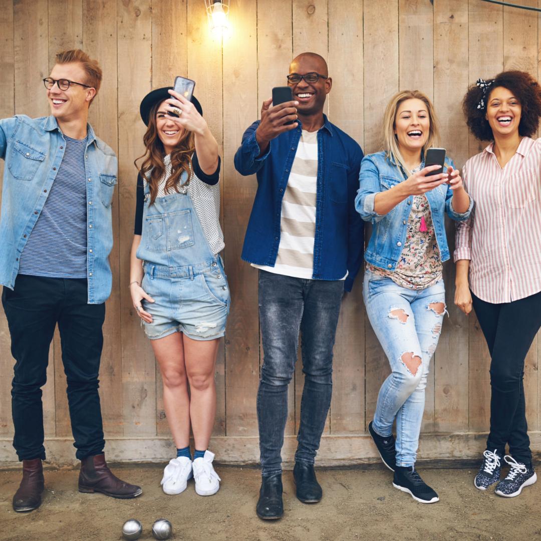 Vijf vrienden lachen en maken selfies met mobiele telefoons tegen een houten achtergrond.