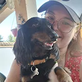 Une femme portant un chapeau bleu et des lunettes sourit en tenant un chien noir avec des taches marron et un collier orange.