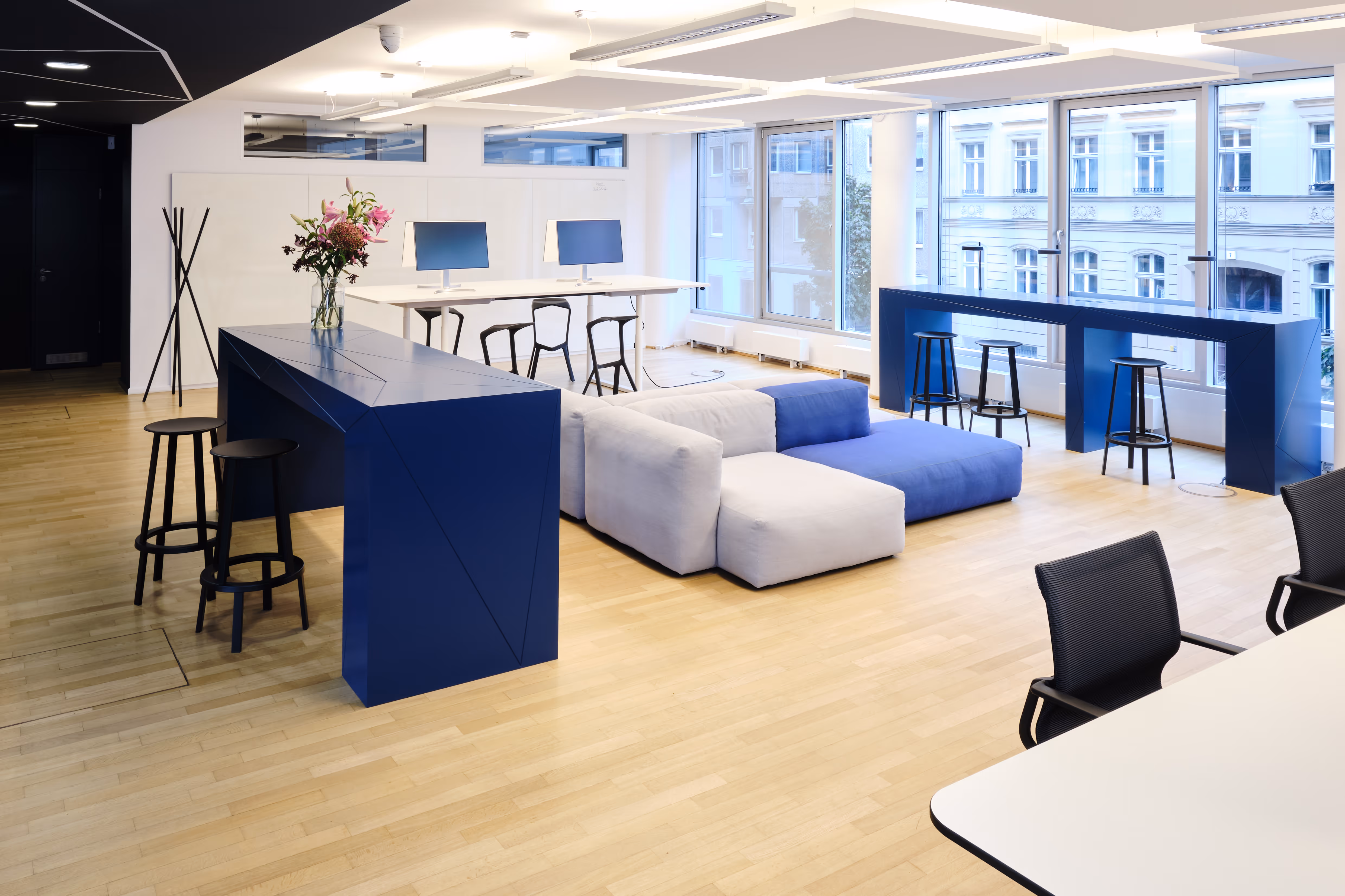 Modernes Büro mit großen Fenstern, blau-weißer Lounge-Couch, blauen Stehtischen mit Barhockern und Arbeitstischen mit Computern.