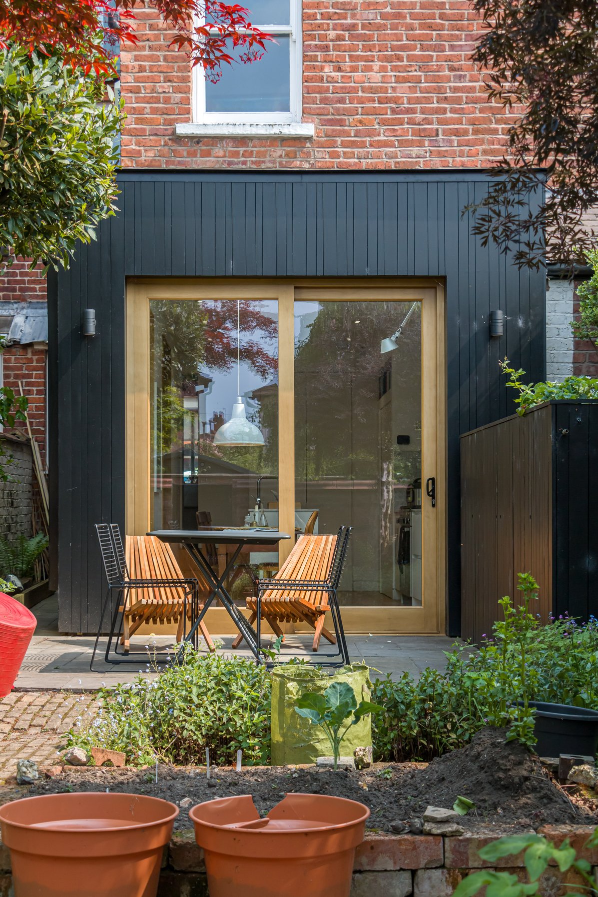 Backyard garden with two wooden chairs, a black table, and sliding glass door opening into a brick house.