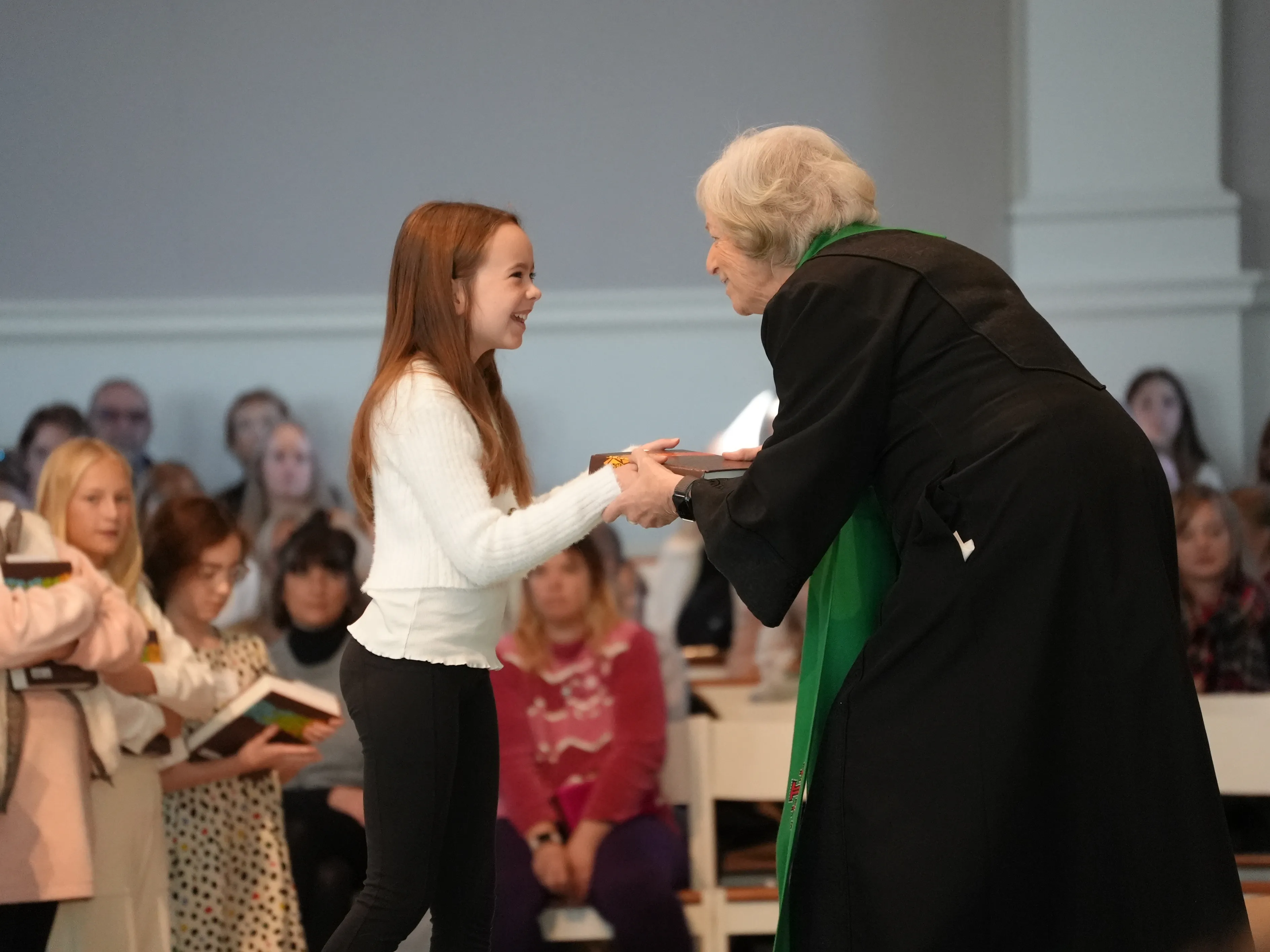 Young girl receiving a book from an elderly woman dressed in black with a green stole, in front of an audience.