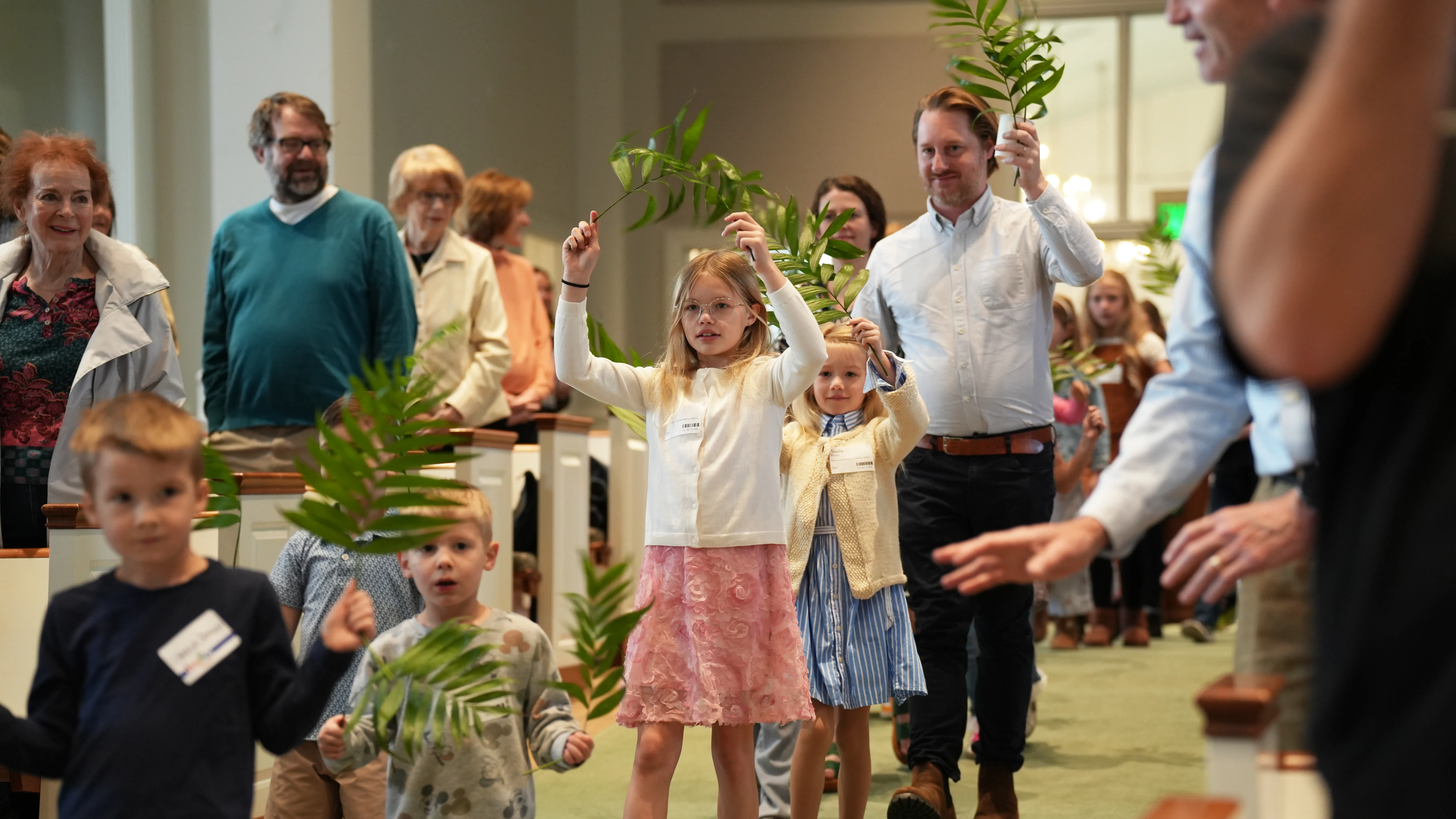 Children and adults walking down a church aisle holding palm branches, likely during a Palm Sunday procession.
