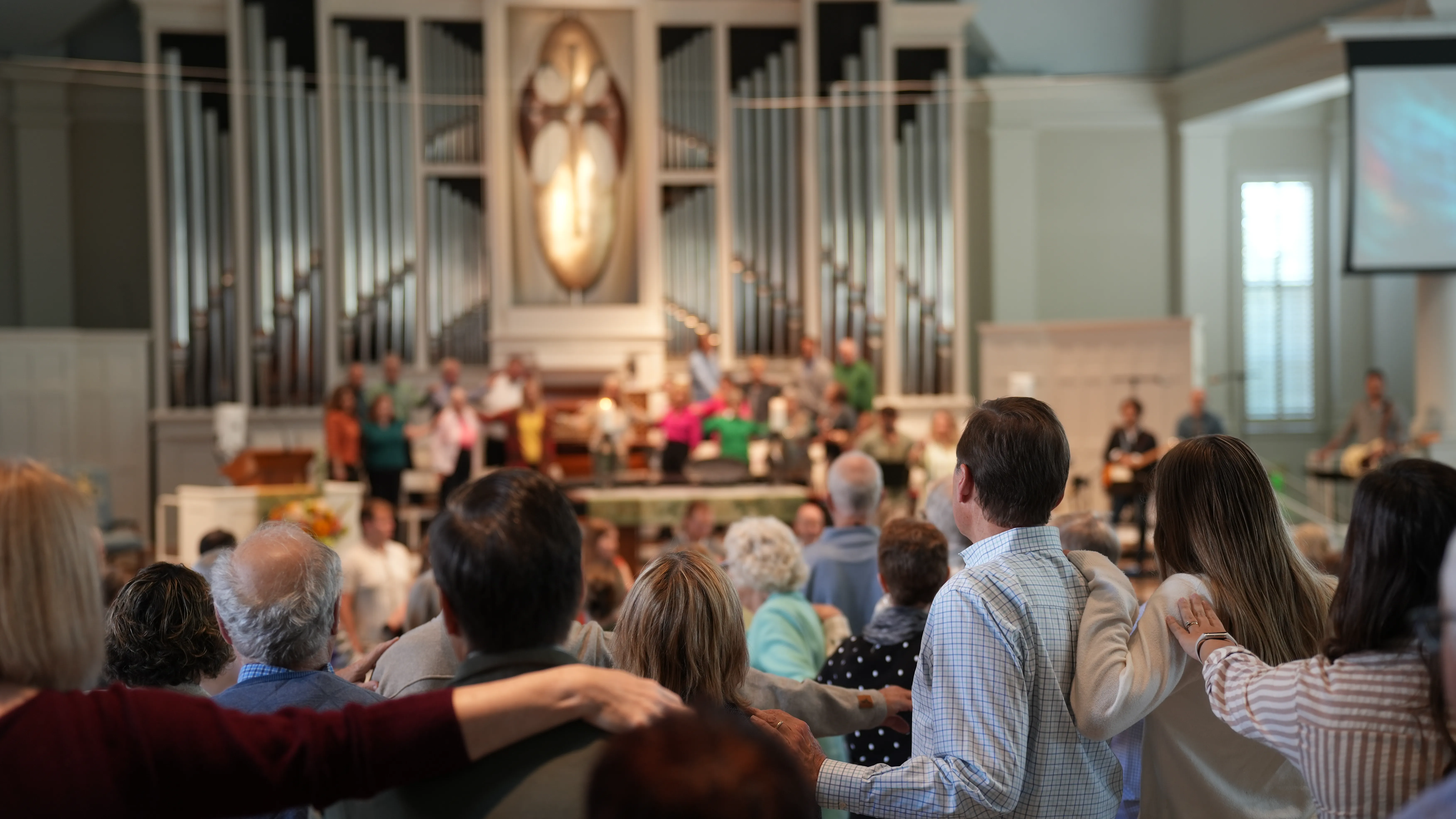 Congregation with linked arms facing a choir and organ pipes inside a church sanctuary.