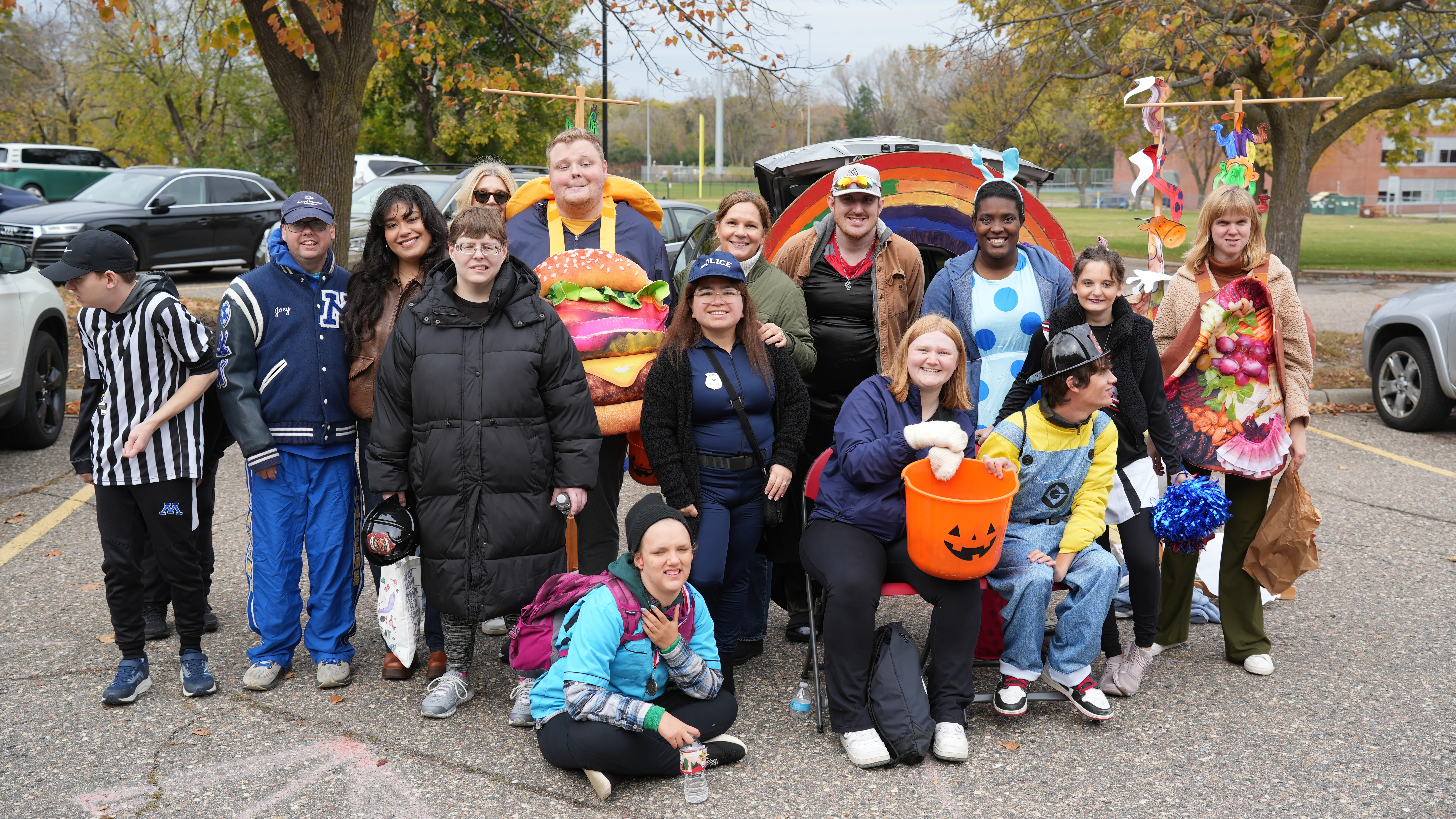 Group of people in various costumes including a hamburger, police officer, minion, and carrot, posing outdoors in a parking lot with autumn trees.