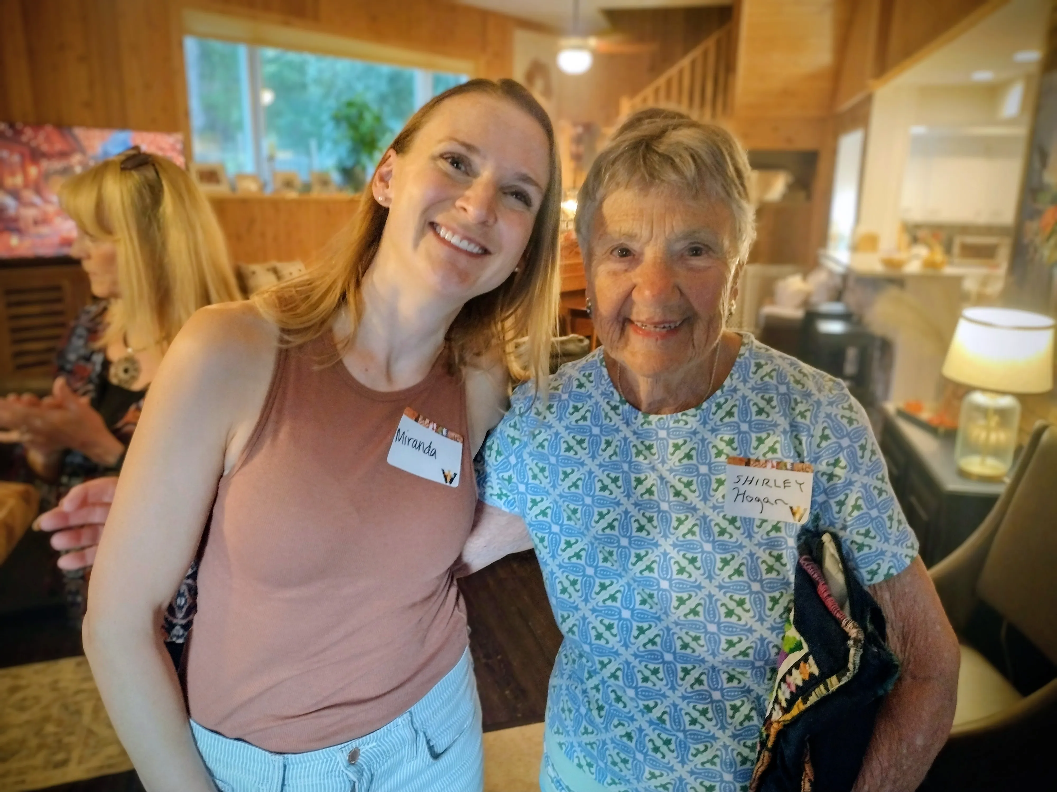 Two smiling women wearing name tags, one younger in a sleeveless top and the other older in a patterned shirt, posing inside a warmly lit home.