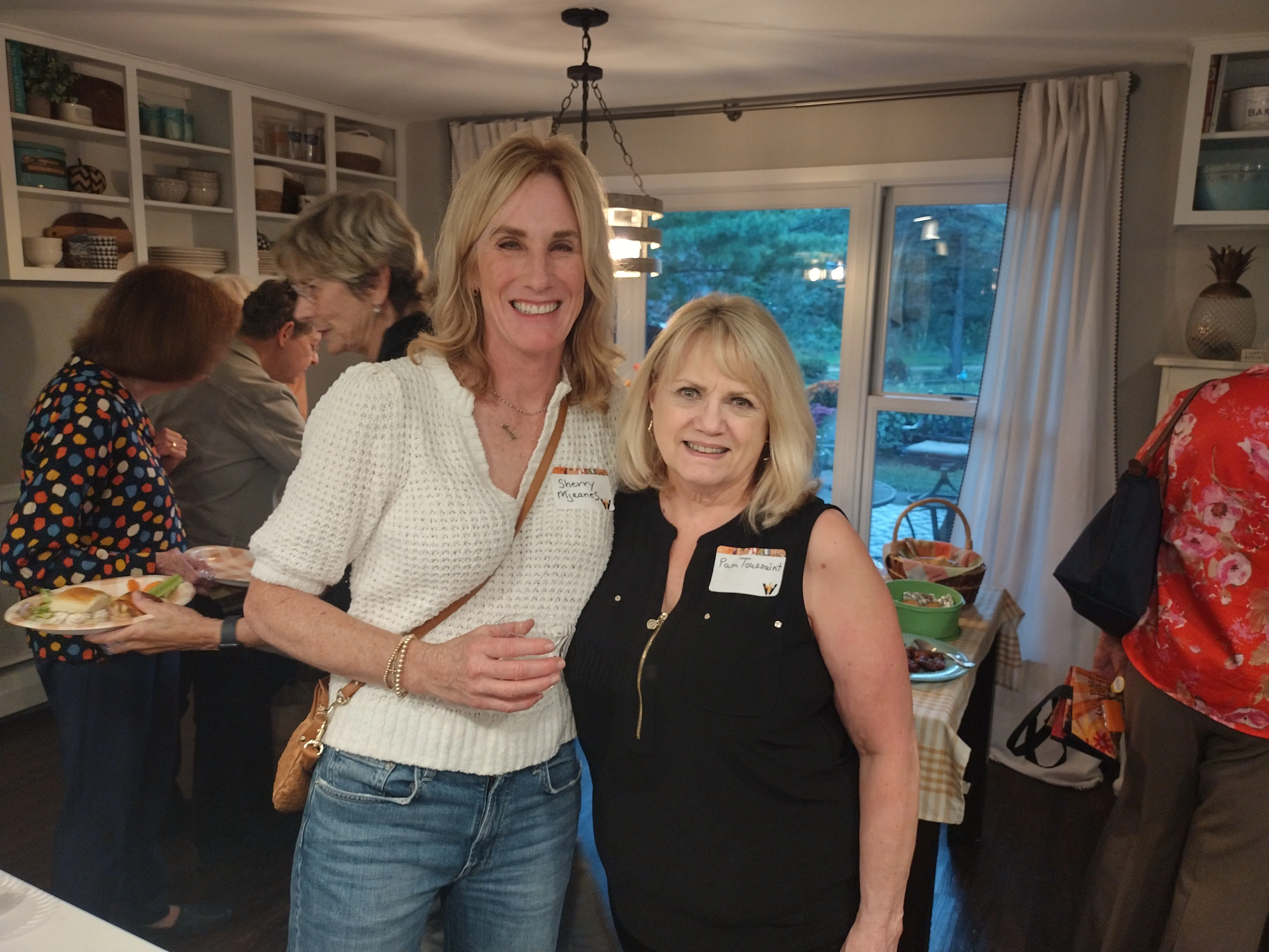 Two smiling women wearing name tags stand together in a kitchen during a social gathering.
