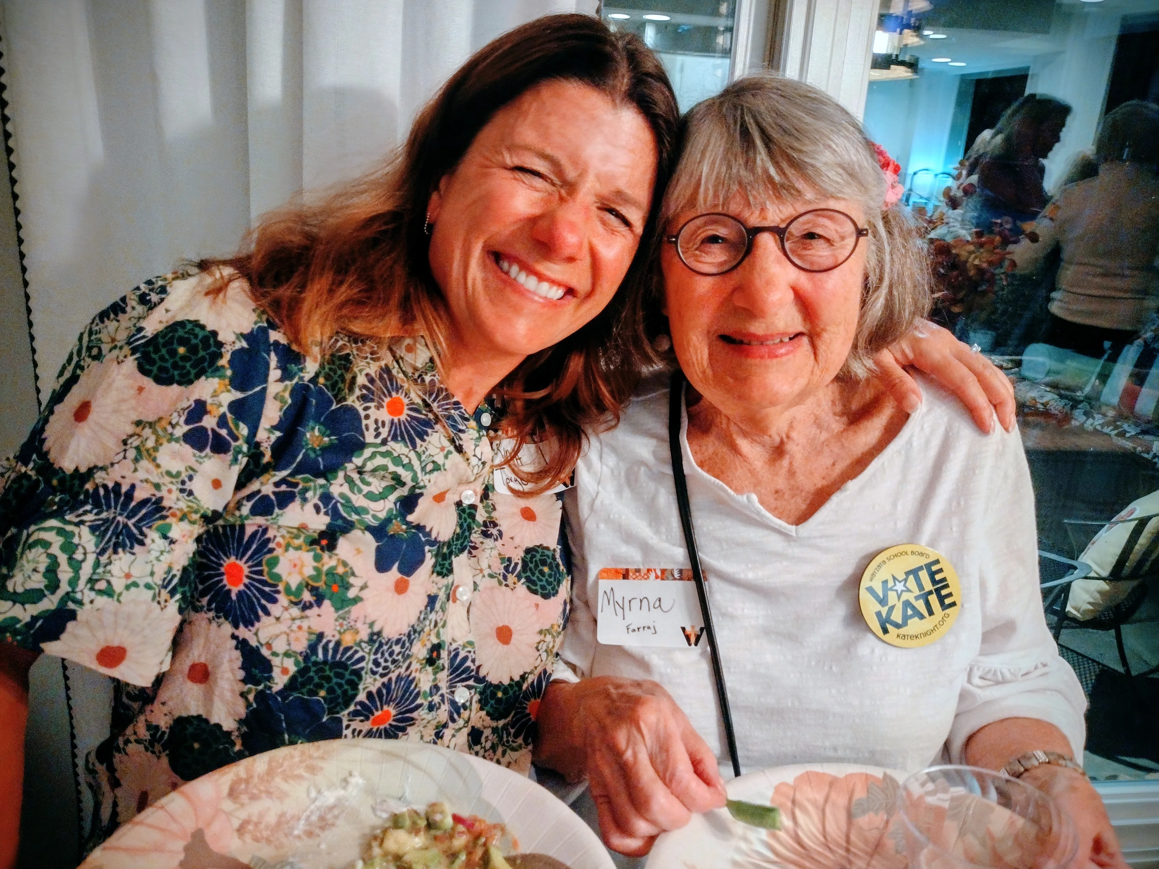 Two women smiling and posing closely together at a gathering, holding plates of food.