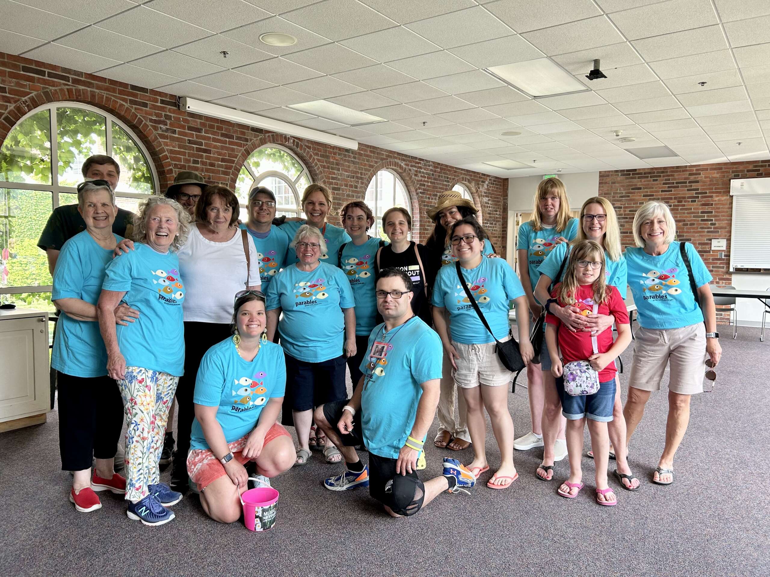 Group of diverse adults and children indoors, most wearing matching blue Parables t-shirts with colorful fish graphic, posing and smiling.