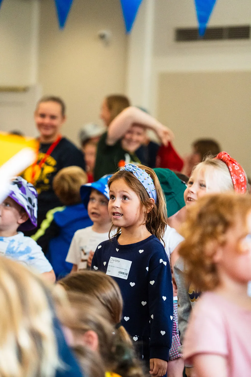 Group of children indoors, some wearing colorful hats and headbands, attentively looking towards something off-camera.