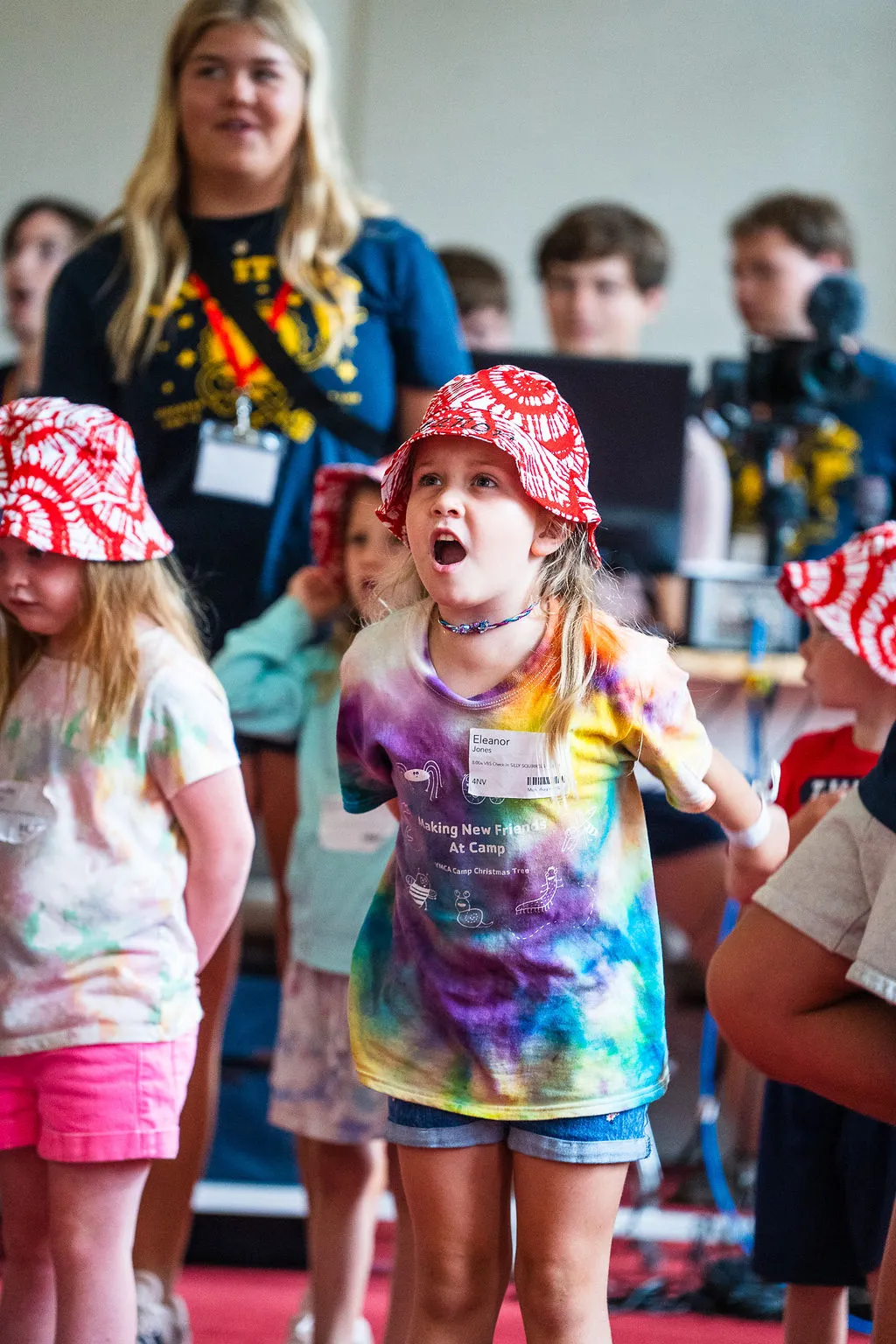 Young girl wearing a red tie-dye bucket hat and a colorful tie-dye shirt with a name tag, speaking or singing with other children and adults around her.