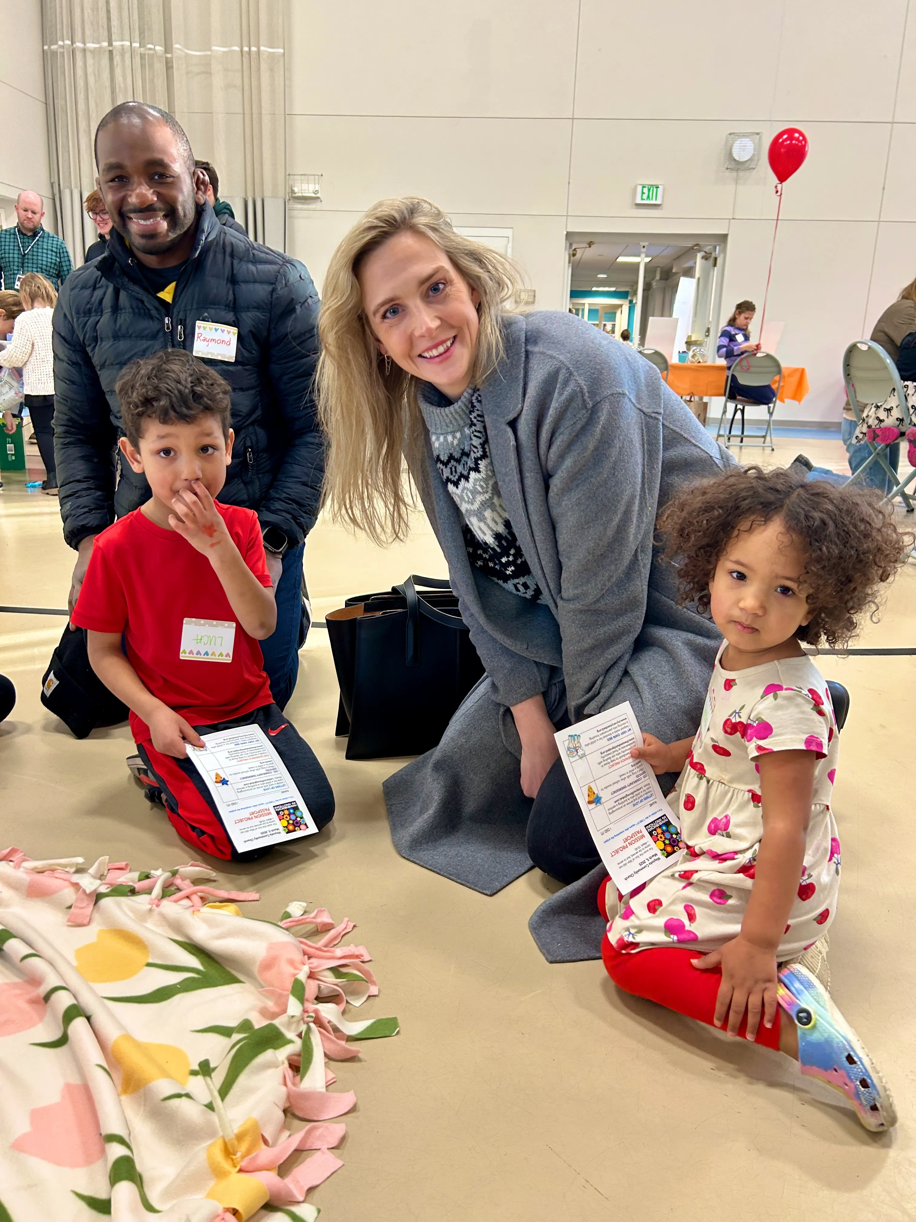 A smiling man and a woman kneeling on the floor with two young children holding event pamphlets in an indoor community space.
