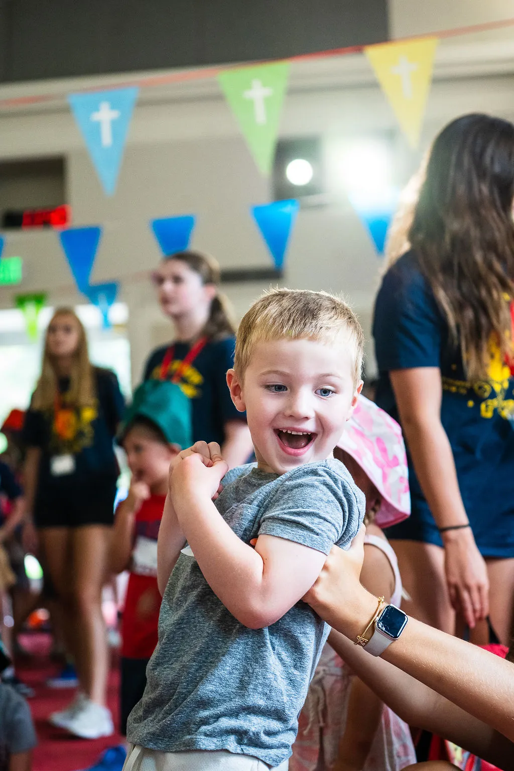 Smiling young boy in a gray shirt being held by an adult with a smartwatch in a lively indoor setting with colorful triangular banners.