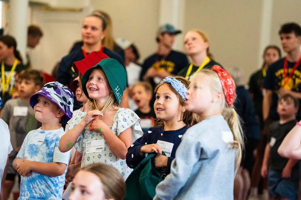 Group of young children indoors, some wearing colorful hats and headbands, looking engaged and smiling.