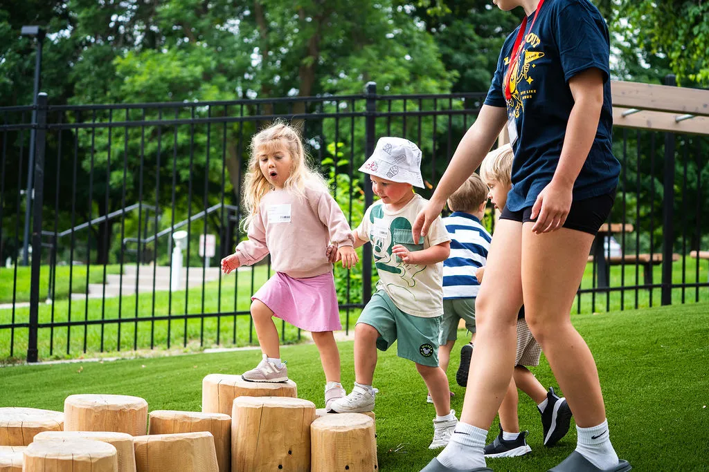 Four children playing outdoors on wooden stumps on grass with a supervising adult nearby.