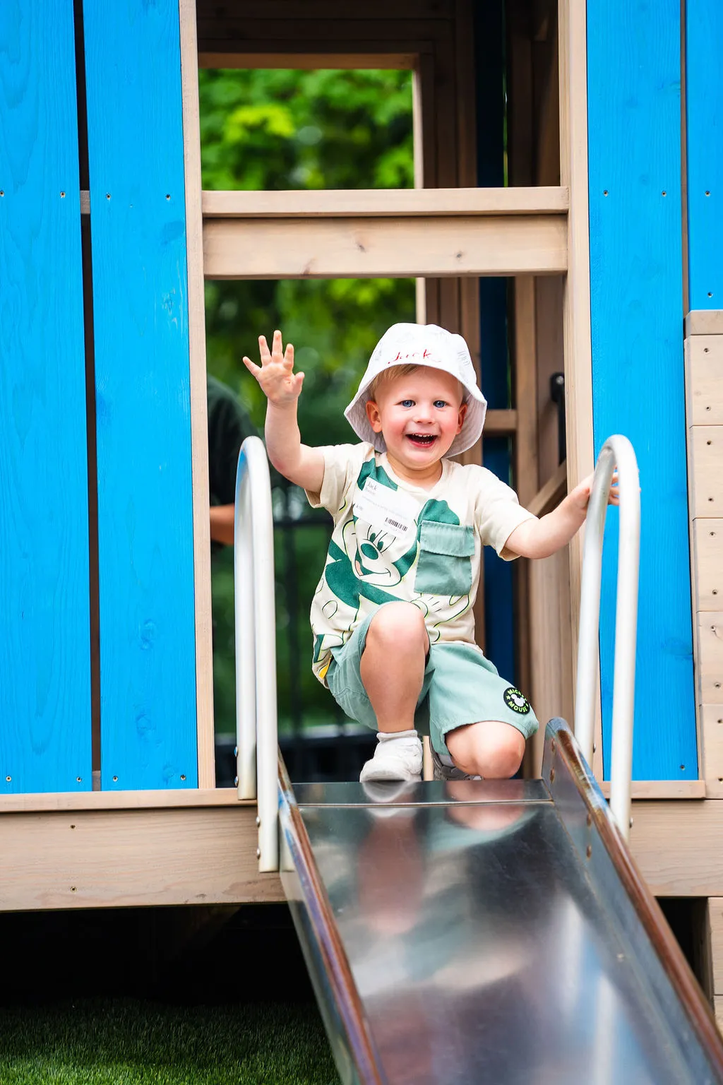 Smiling toddler wearing a white hat and Mickey Mouse shirt sliding down a metal slide at a playground.