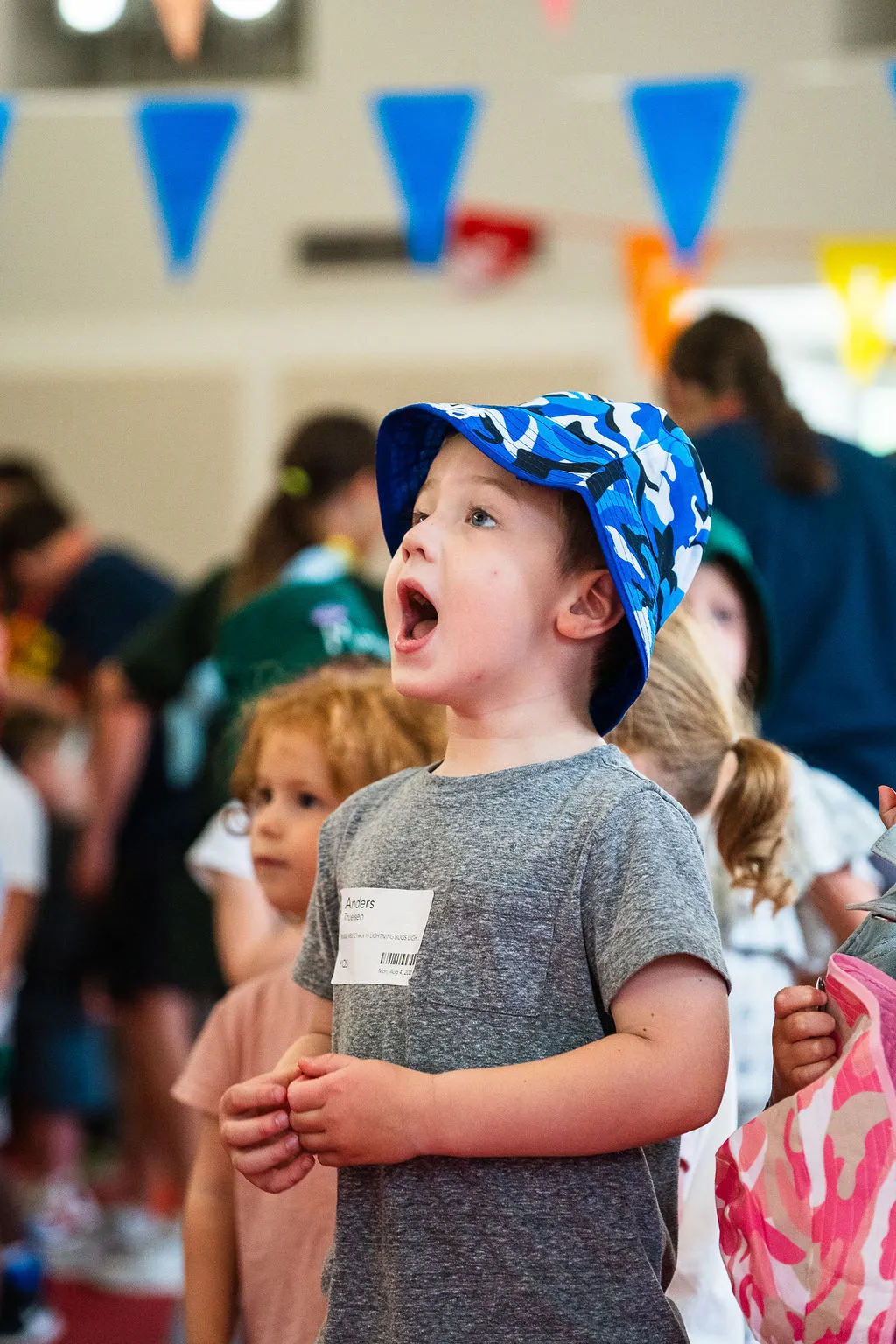 Young boy wearing a blue camouflage bucket hat and gray shirt looking up with mouth open in a busy indoor setting with other children in background.
