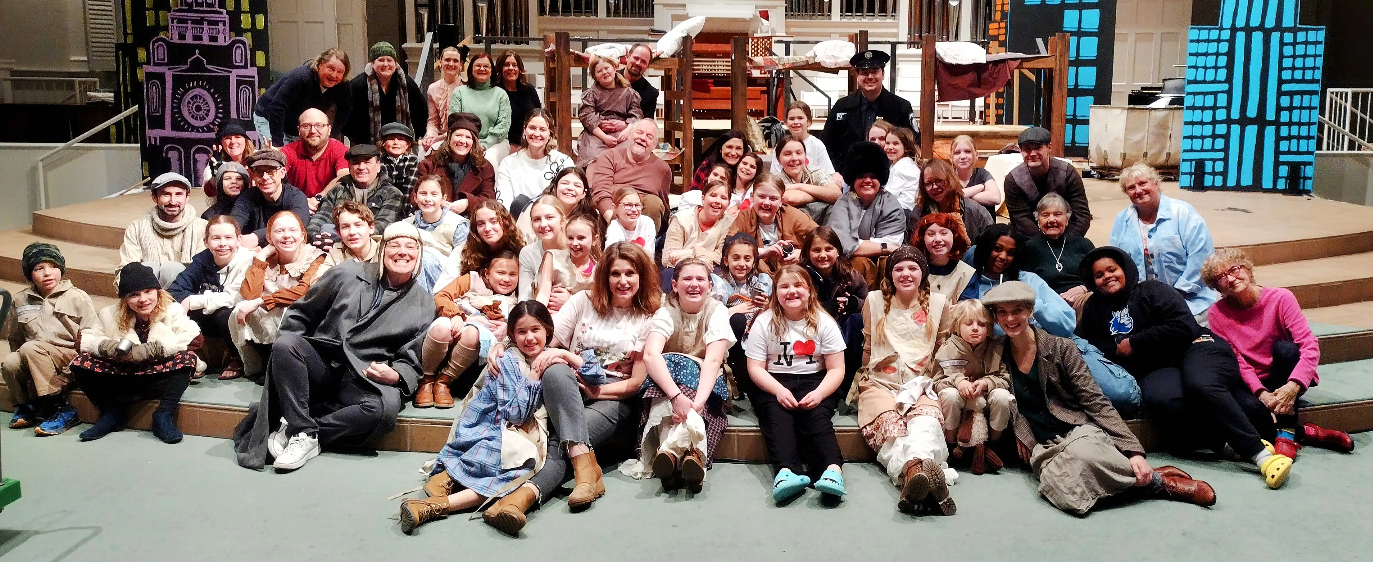 Large diverse group of people of various ages seated and standing on stage in costume, posing for a group photo for a theatrical production.
