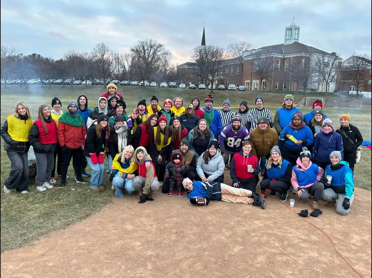 Large group of people outdoors on a field wearing colored pinnies and winter clothing, posing together near a baseball diamond with buildings and cars in the background.