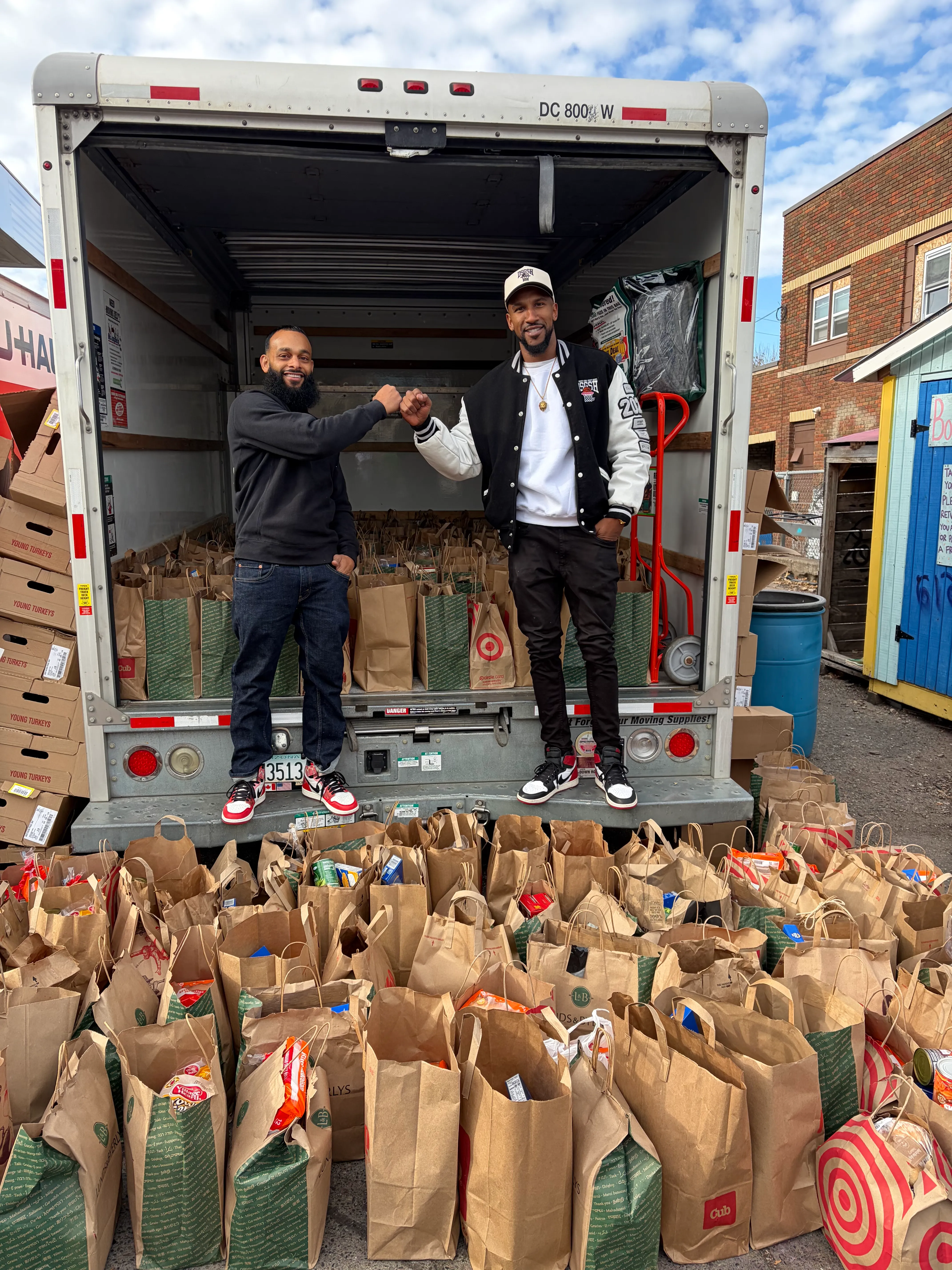 Two men fist bumping while standing on the back of a delivery truck filled with grocery bags, with more grocery bags spread out on the ground in front of the truck.
