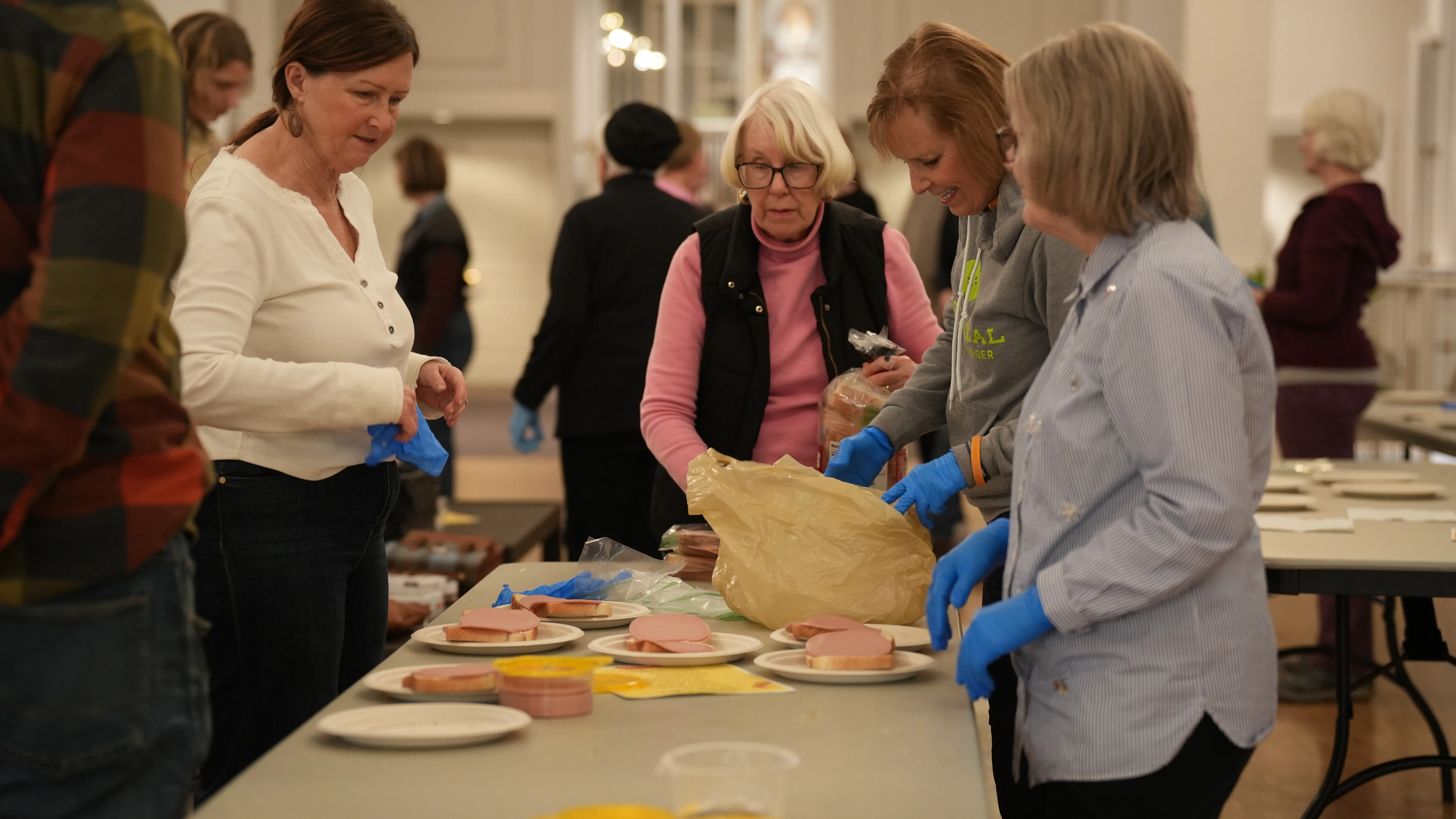 Volunteers wearing blue gloves assembling sandwiches on paper plates at a community event in a large room.