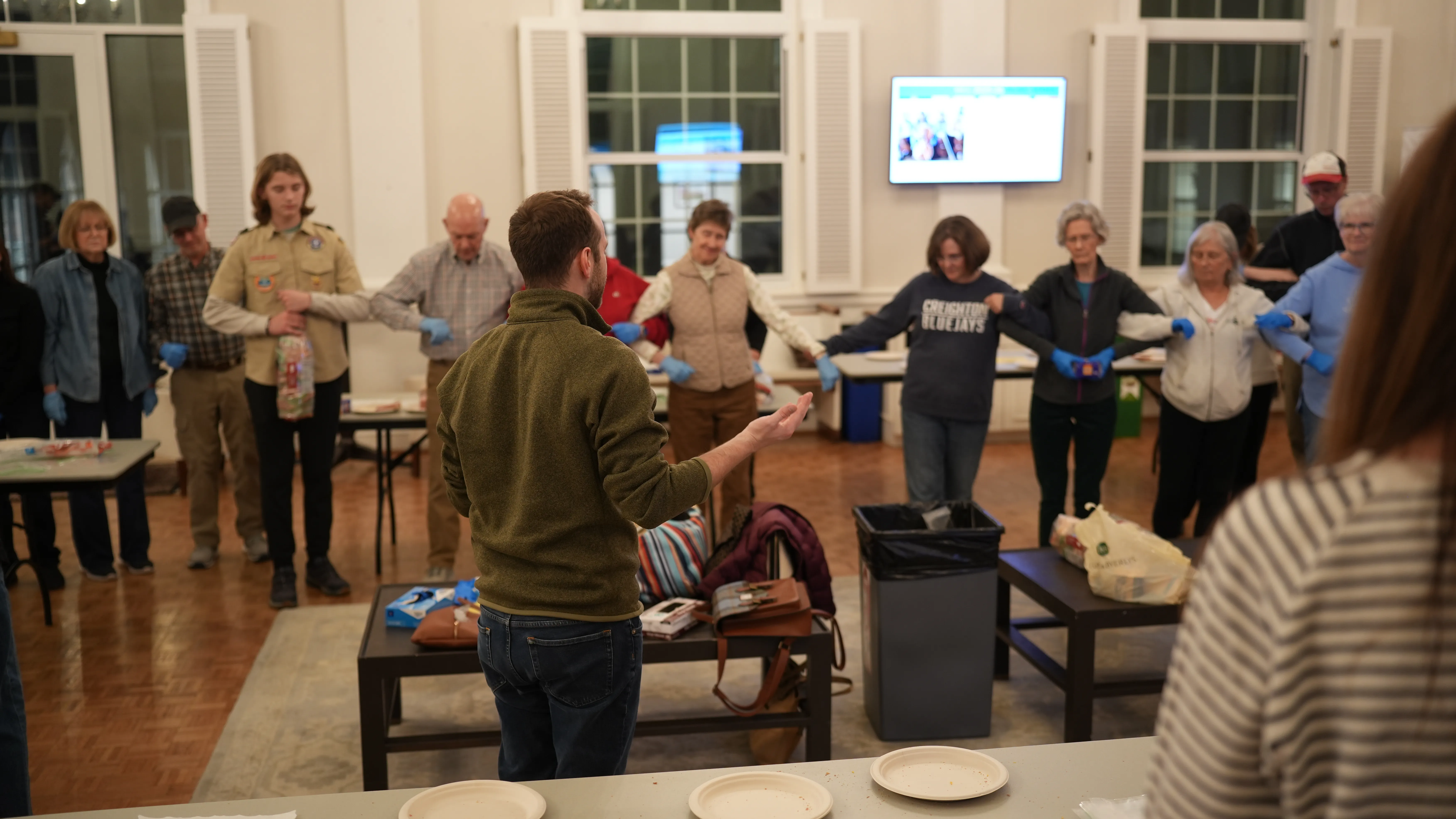 Group of people standing in a circle holding hands, indoors with a man addressing them from the center.