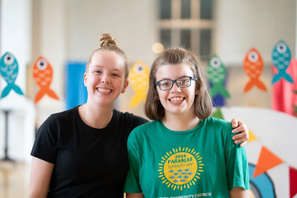 Two smiling young women standing side by side, one with glasses and a green '2024 Parables Summer Camp' shirt, with colorful fish decorations in the background.