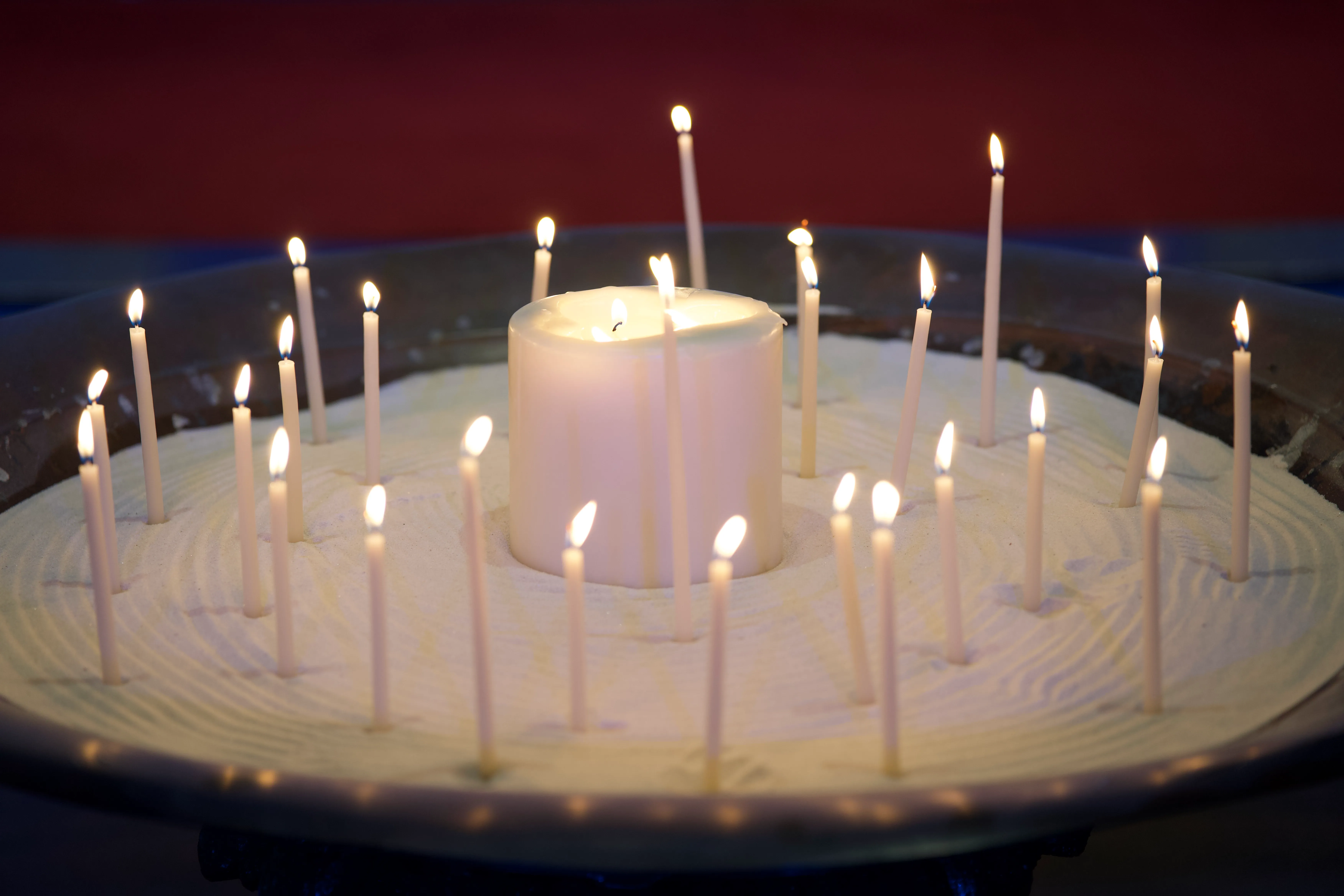 Large white candle surrounded by multiple smaller thin white candles lit and placed in white sand within a round shallow container.