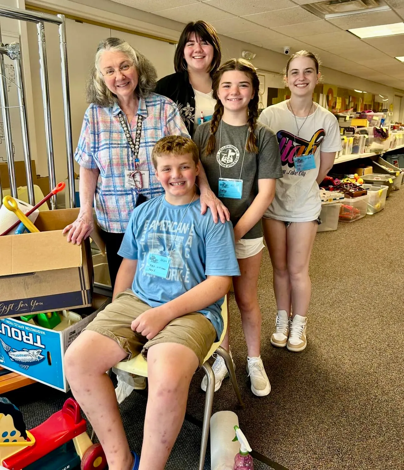 Group of five smiling people posing indoors with shelves of toys and supplies behind them.