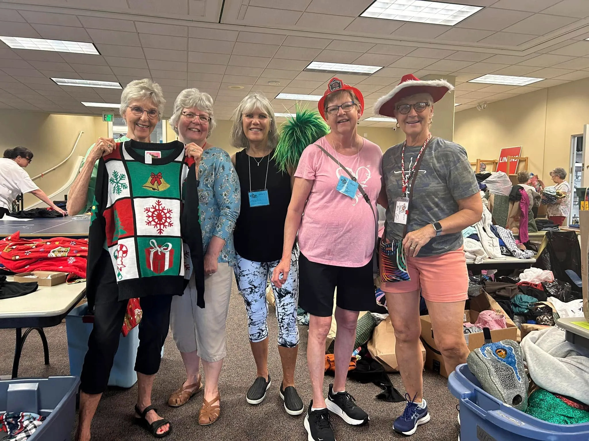 Group of five smiling women standing indoors at a rummage sale, one holding a holiday-themed sweater.