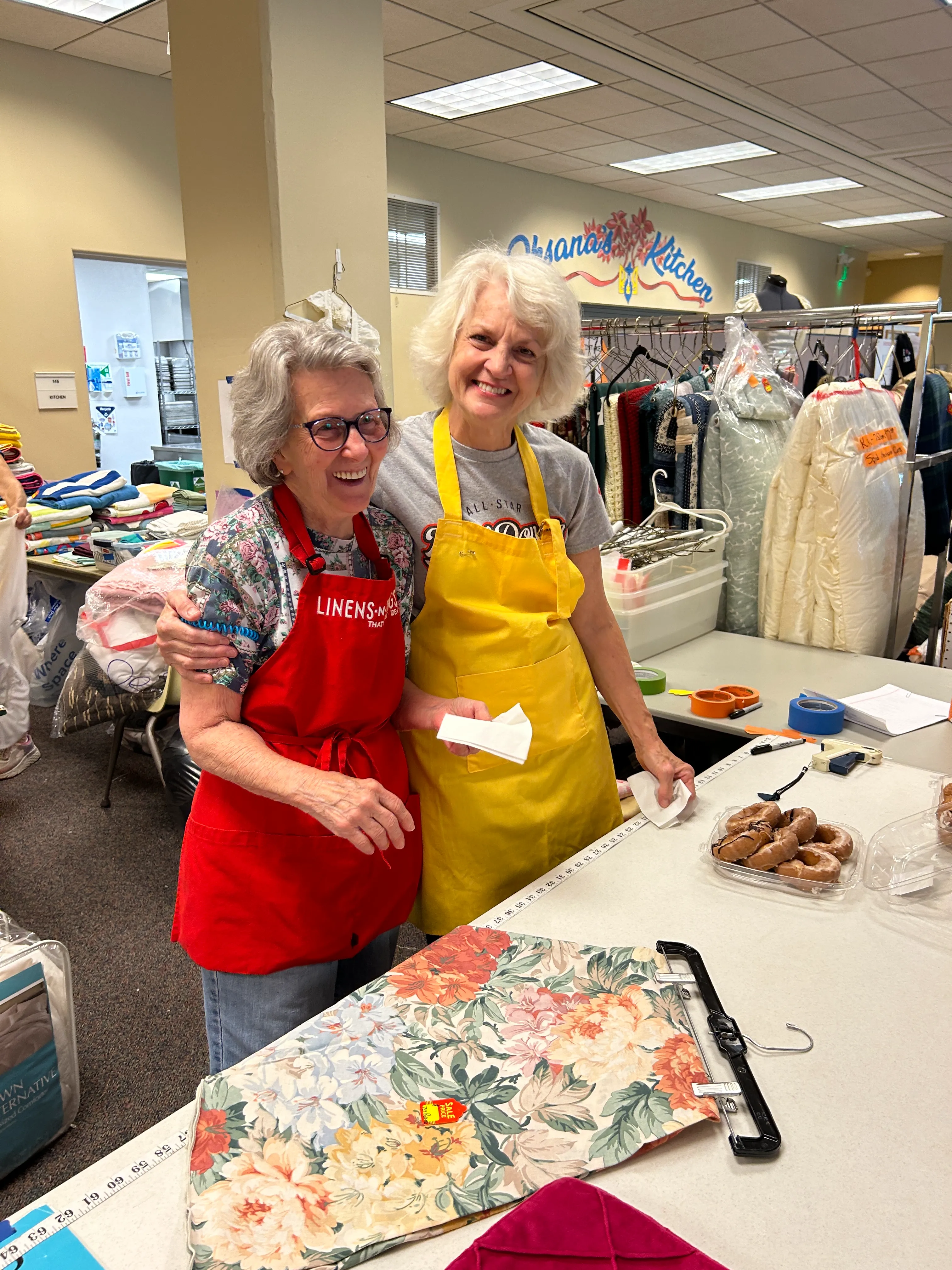 Two smiling elderly women wearing aprons, standing together behind a table with fabric and donuts in a craft or thrift store setting.