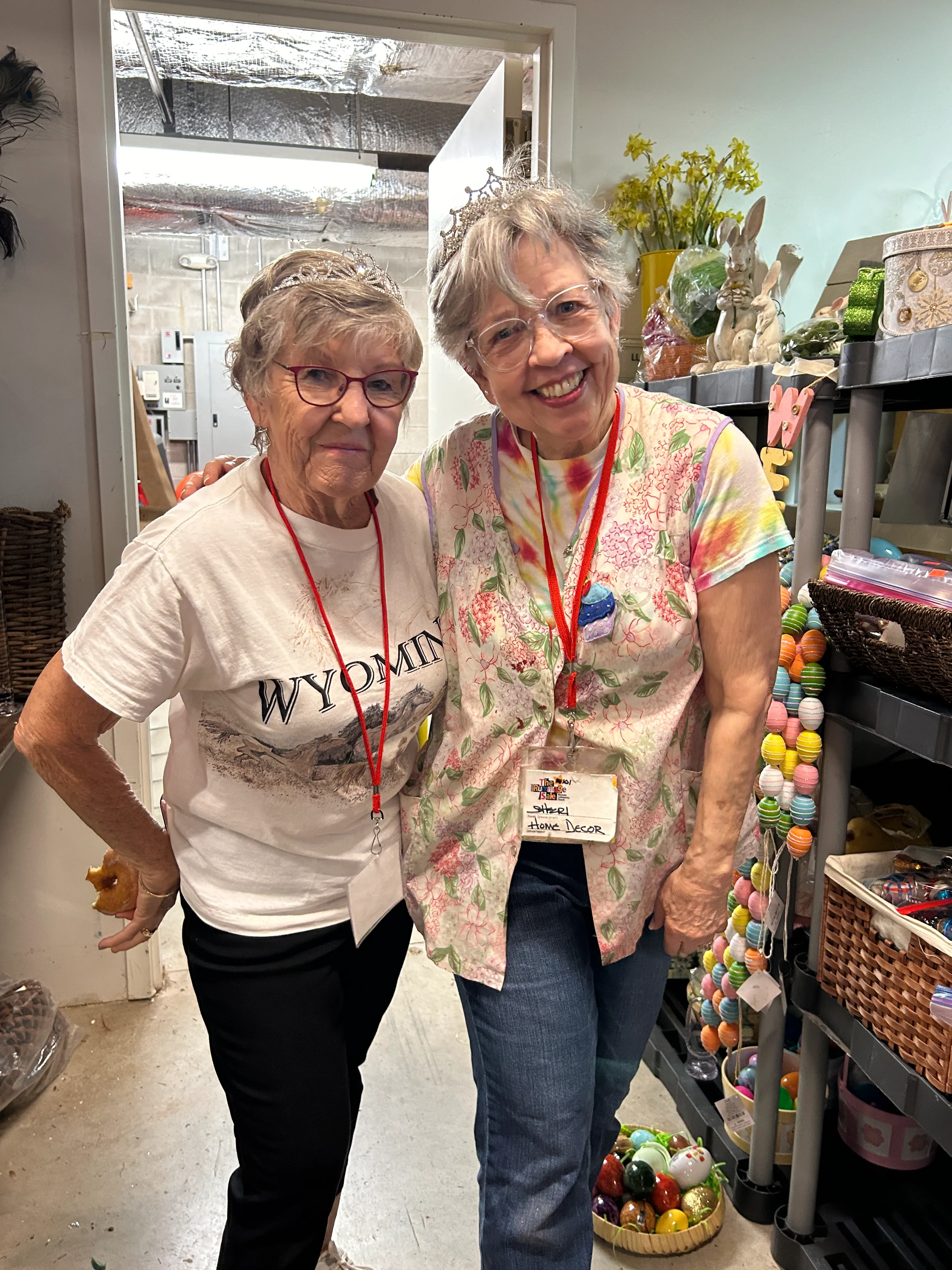 Two elderly women smiling, wearing tiaras and red lanyards, standing close together in a room decorated with colorful Easter eggs and flowers.