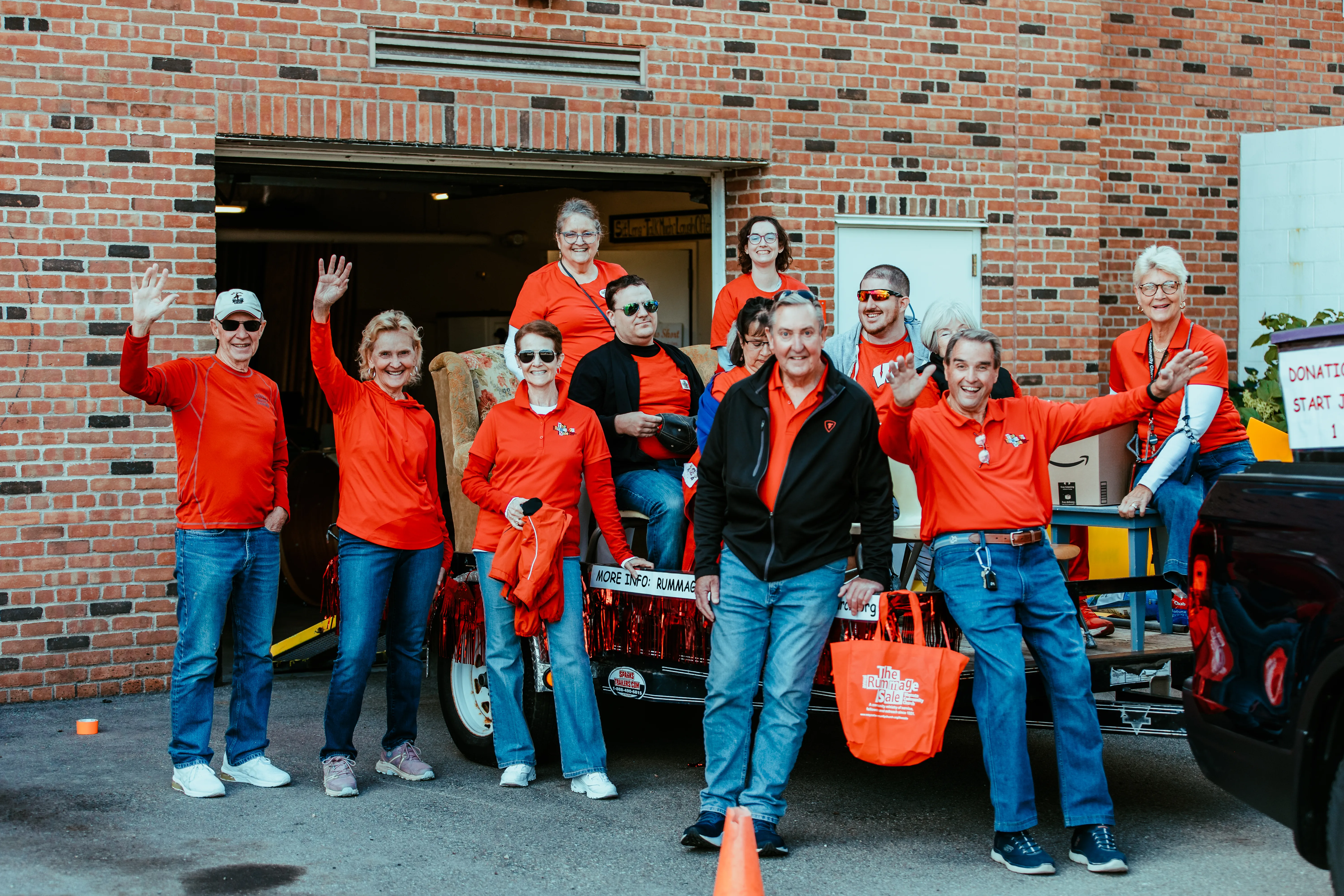Group of smiling people in red shirts waving and posing outdoors near a brick building and trailer.