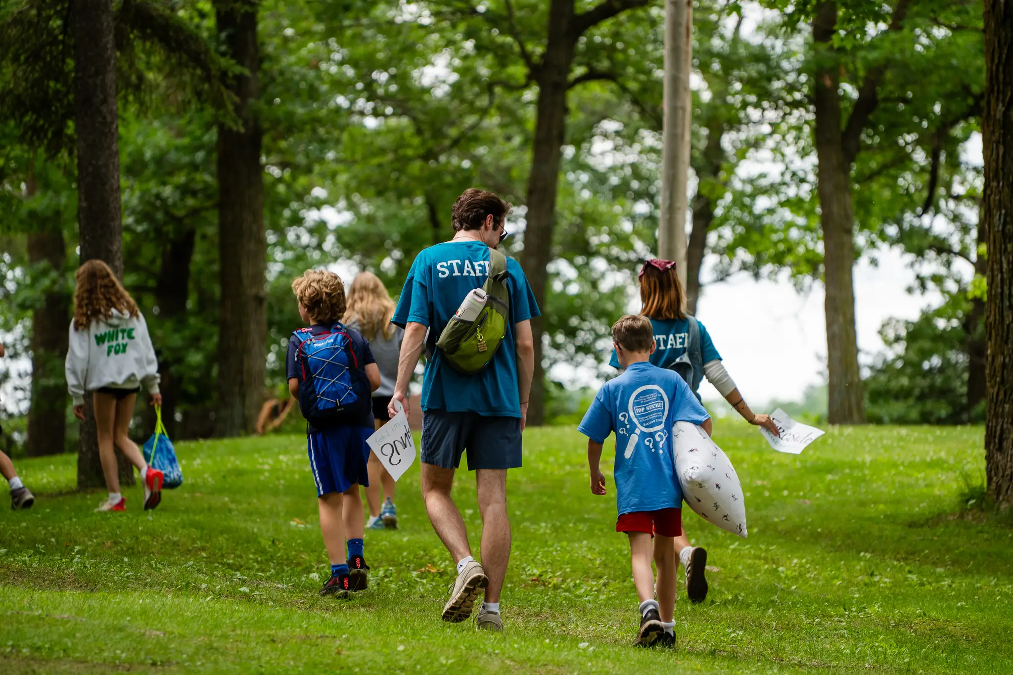 Group of children and two staff members walking on grass in a wooded park during daytime.
