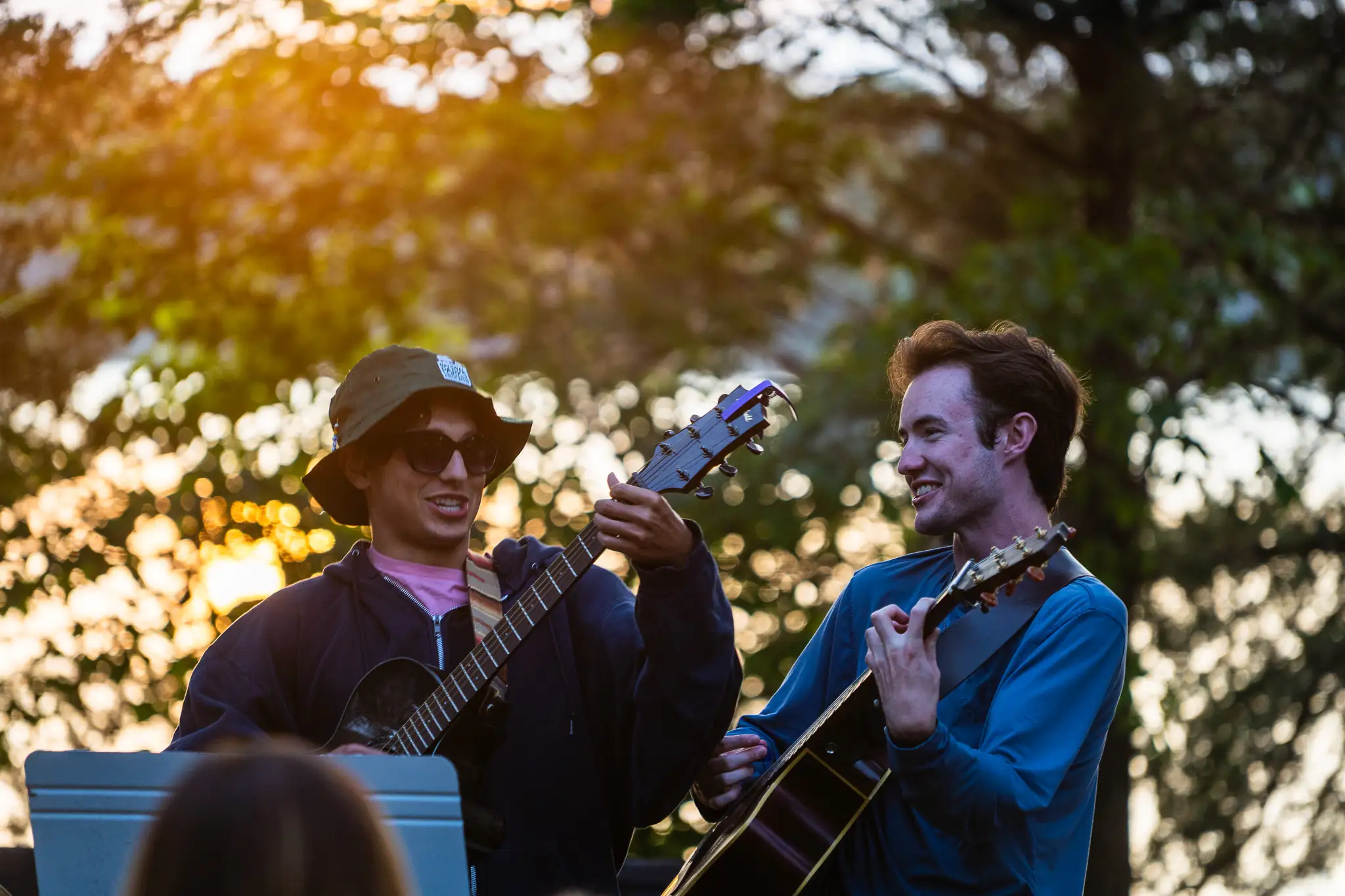 Two young men playing acoustic guitars outdoors at sunset with trees in the background.
