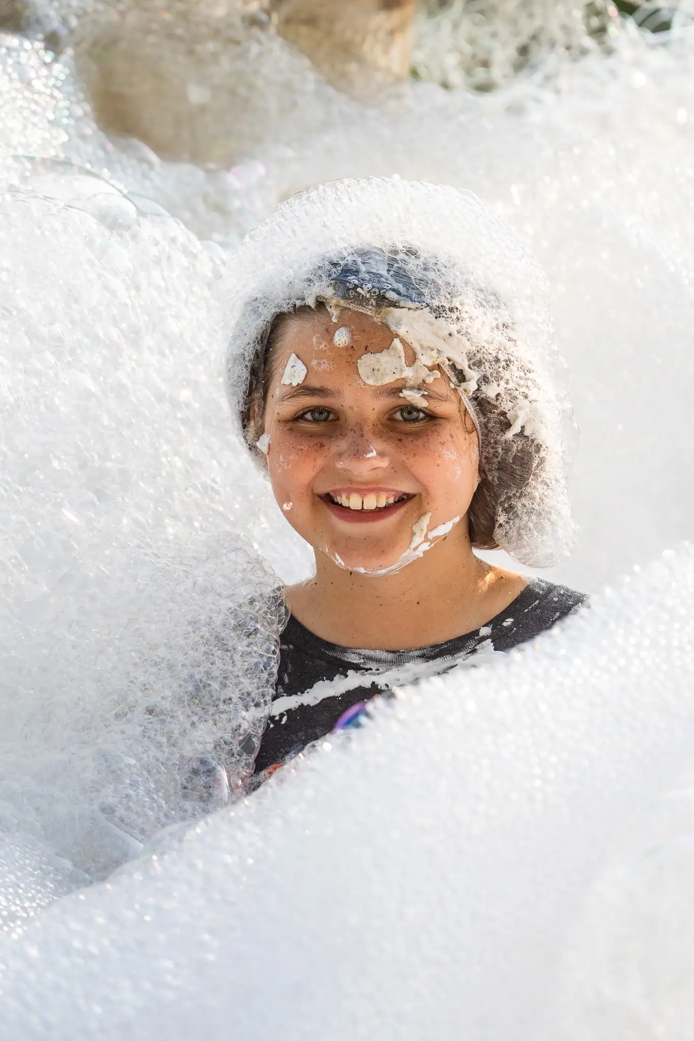 Smiling child covered in soap foam and bubbles while playing in a foam party.