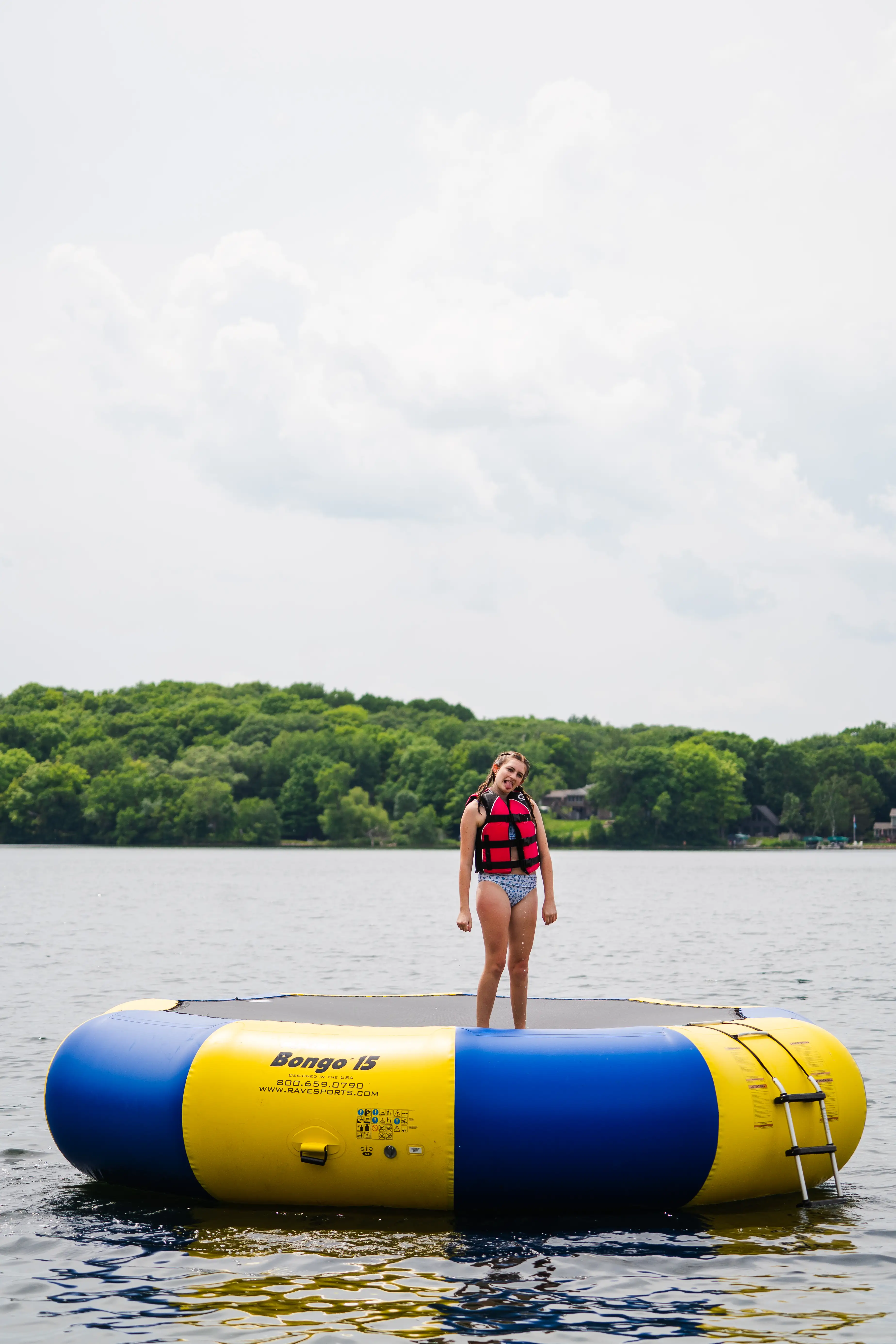 Girl wearing a red life jacket standing on a large yellow and blue inflatable water trampoline on a lake with green trees in the background.