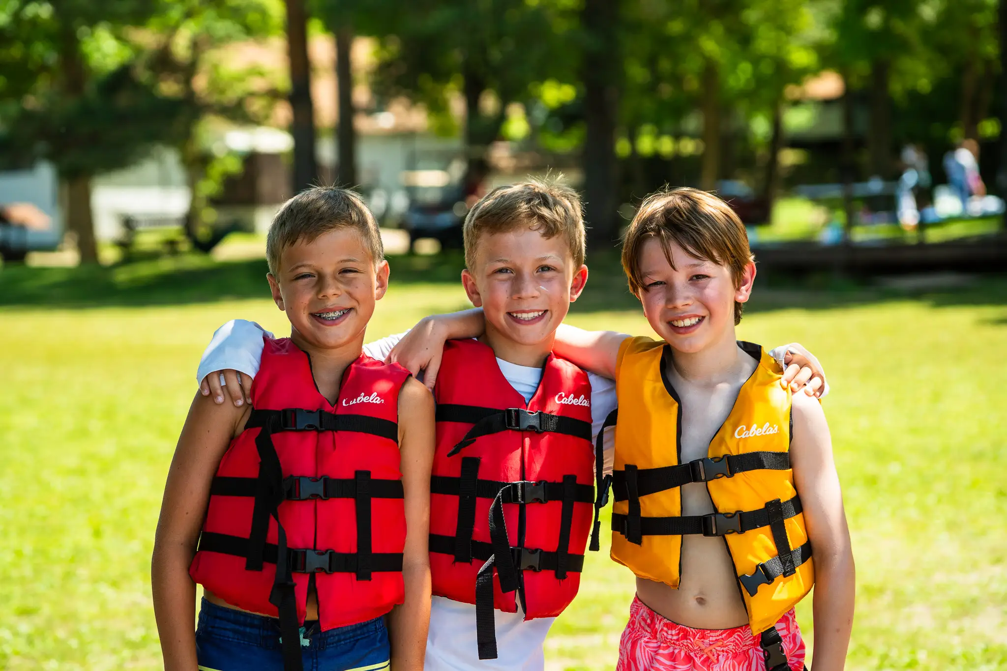 Three smiling boys wearing life jackets with arms around each other on a sunny grassy field.
