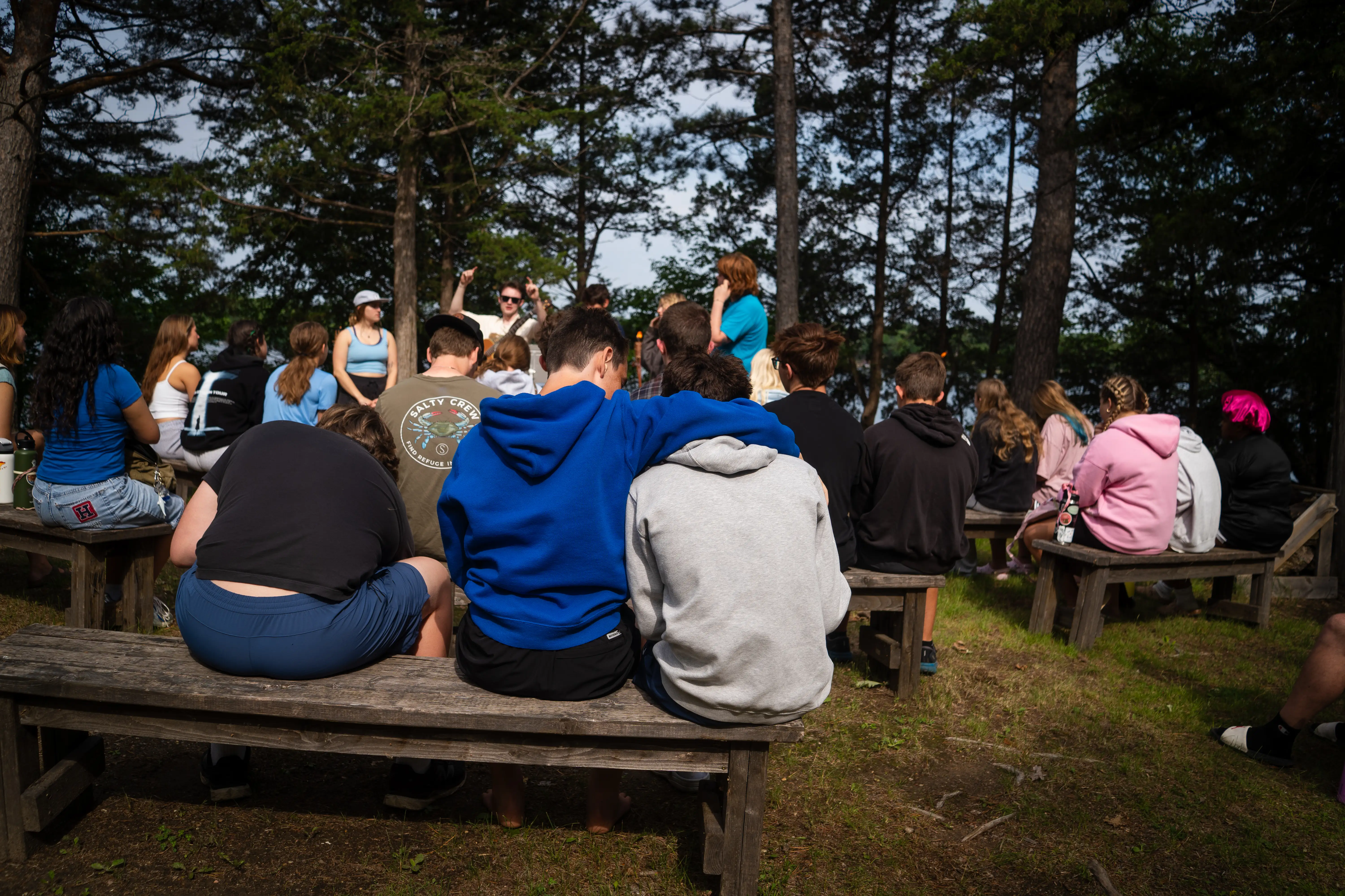 Group of teenagers sitting on wooden benches in a forest clearing, listening to a person speaking in front of them.