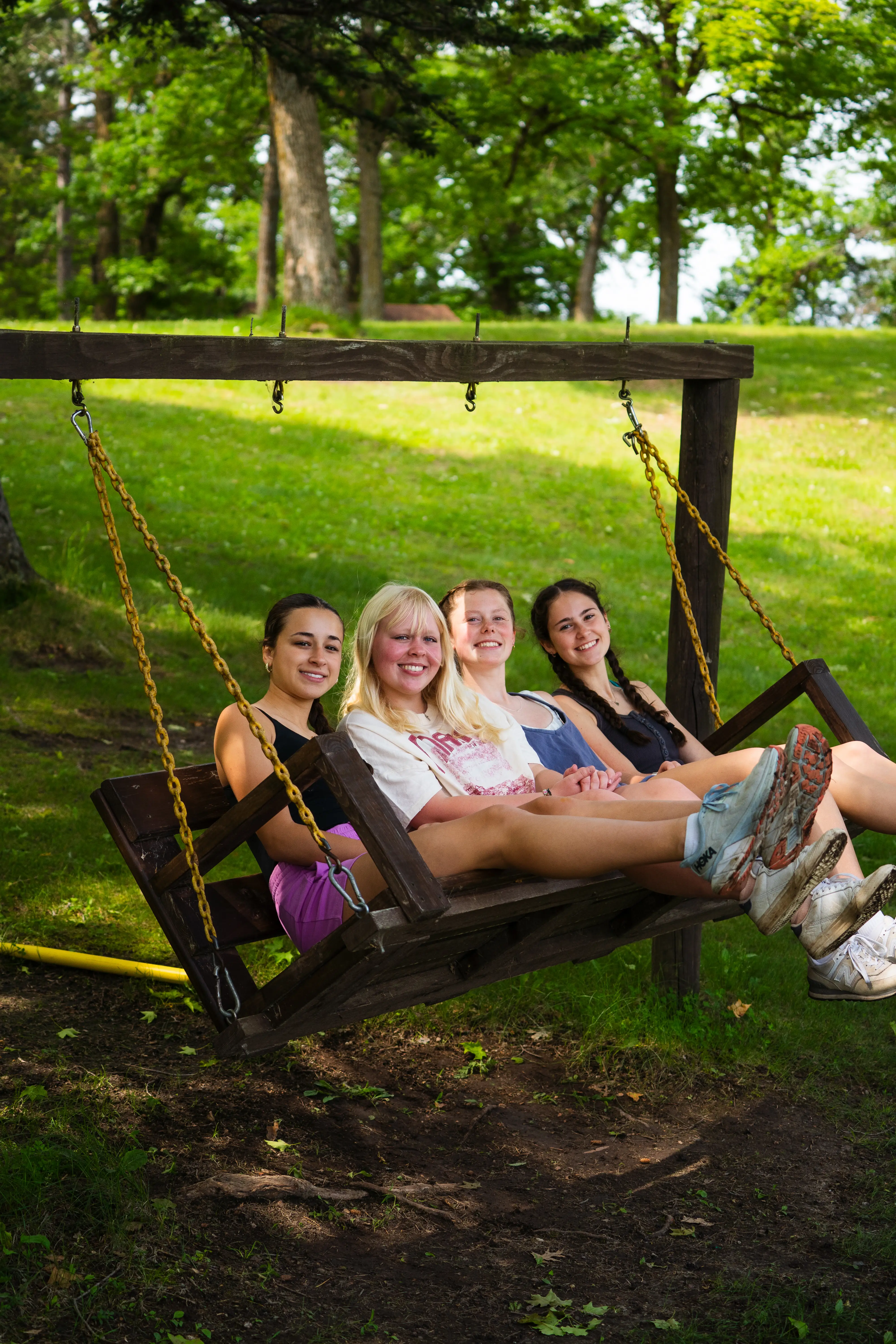 Four young women sitting together on a wooden swing in a park with green grass and trees.