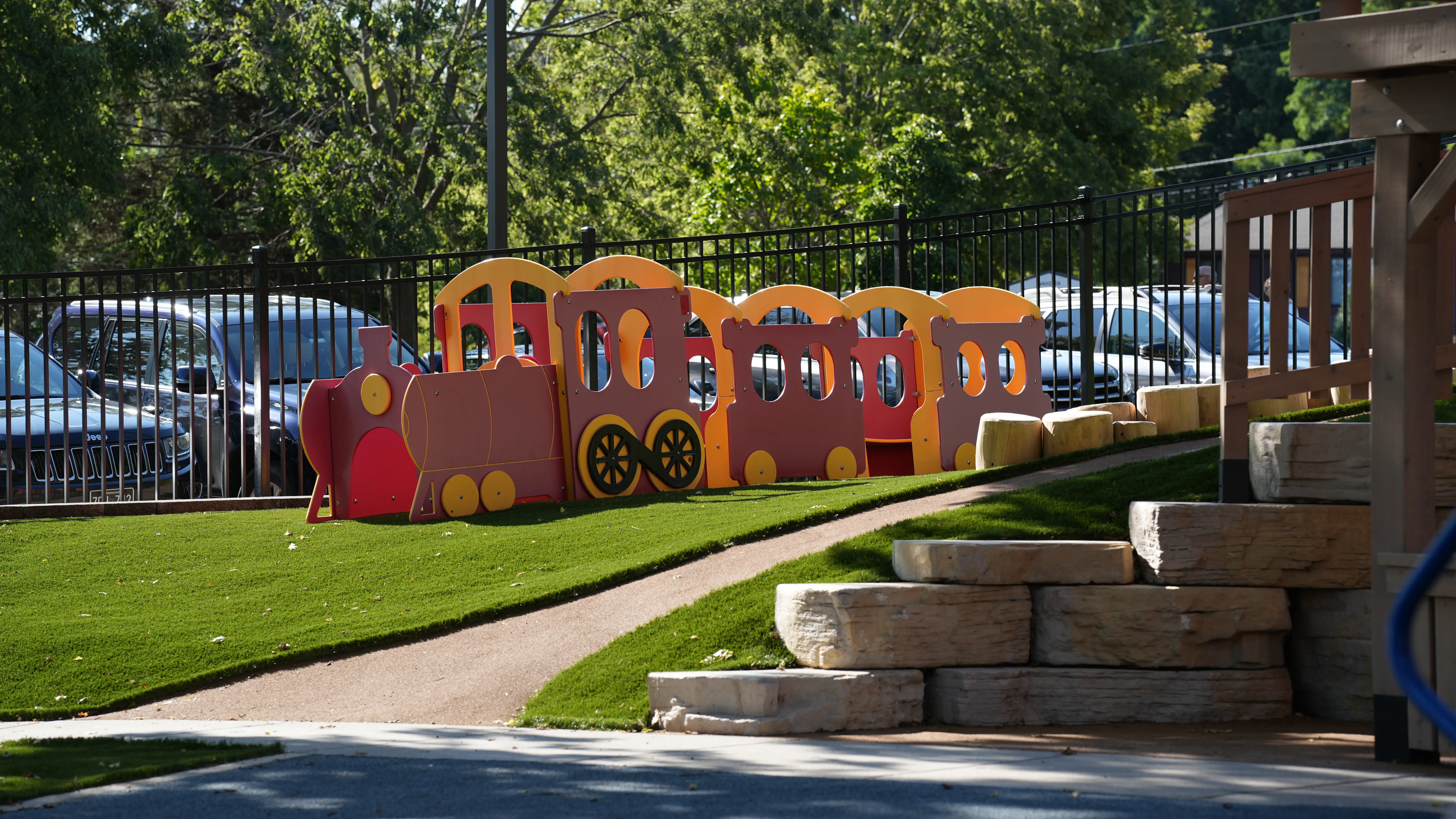 Colorful wooden toy train on artificial grass in an outdoor playground with parked cars and trees in the background.