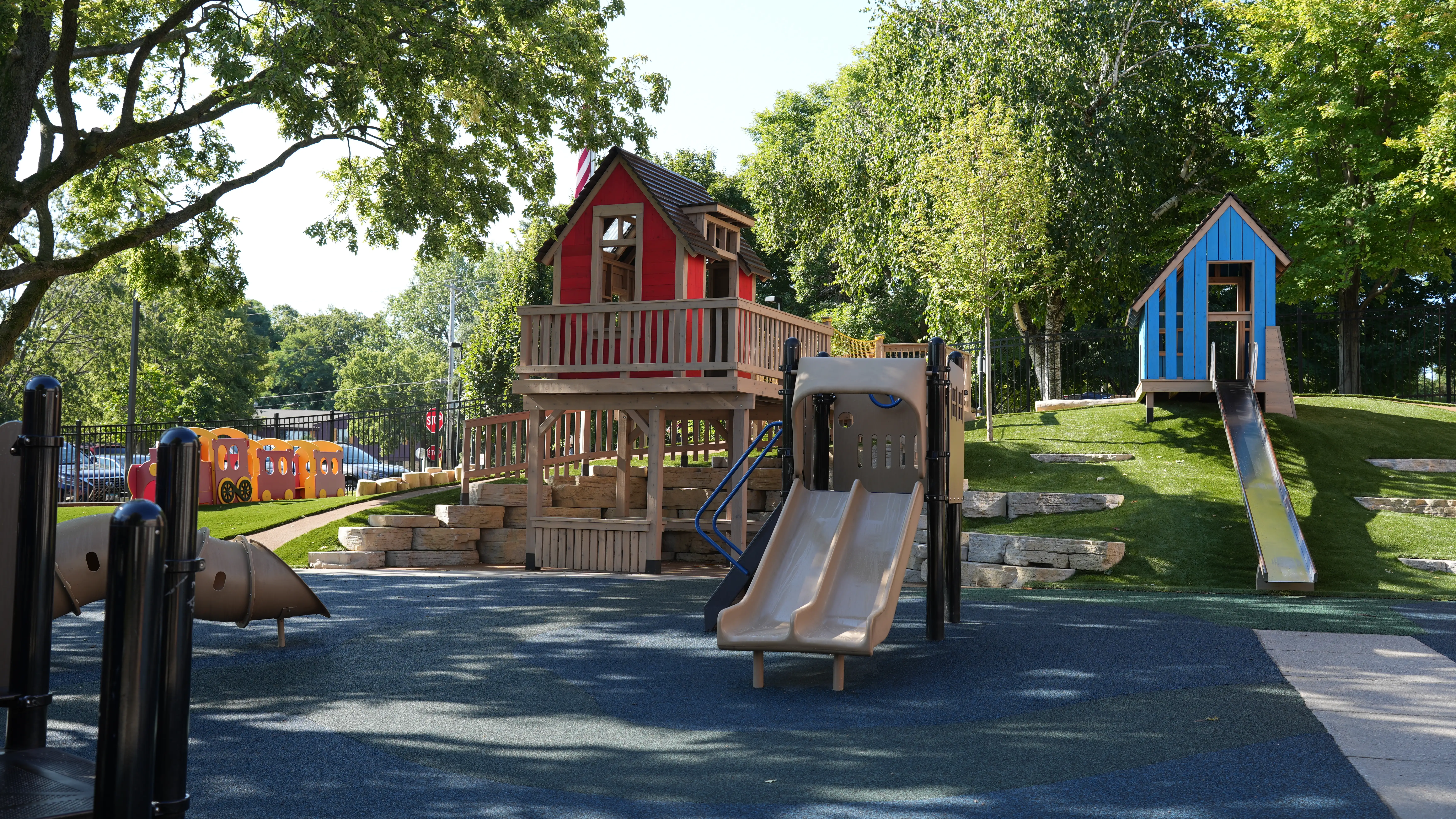 Playground with a double beige slide, a red wooden playhouse on stilts, a blue wooden playhouse with a metal slide, and a plastic train structure, surrounded by green trees and grass.